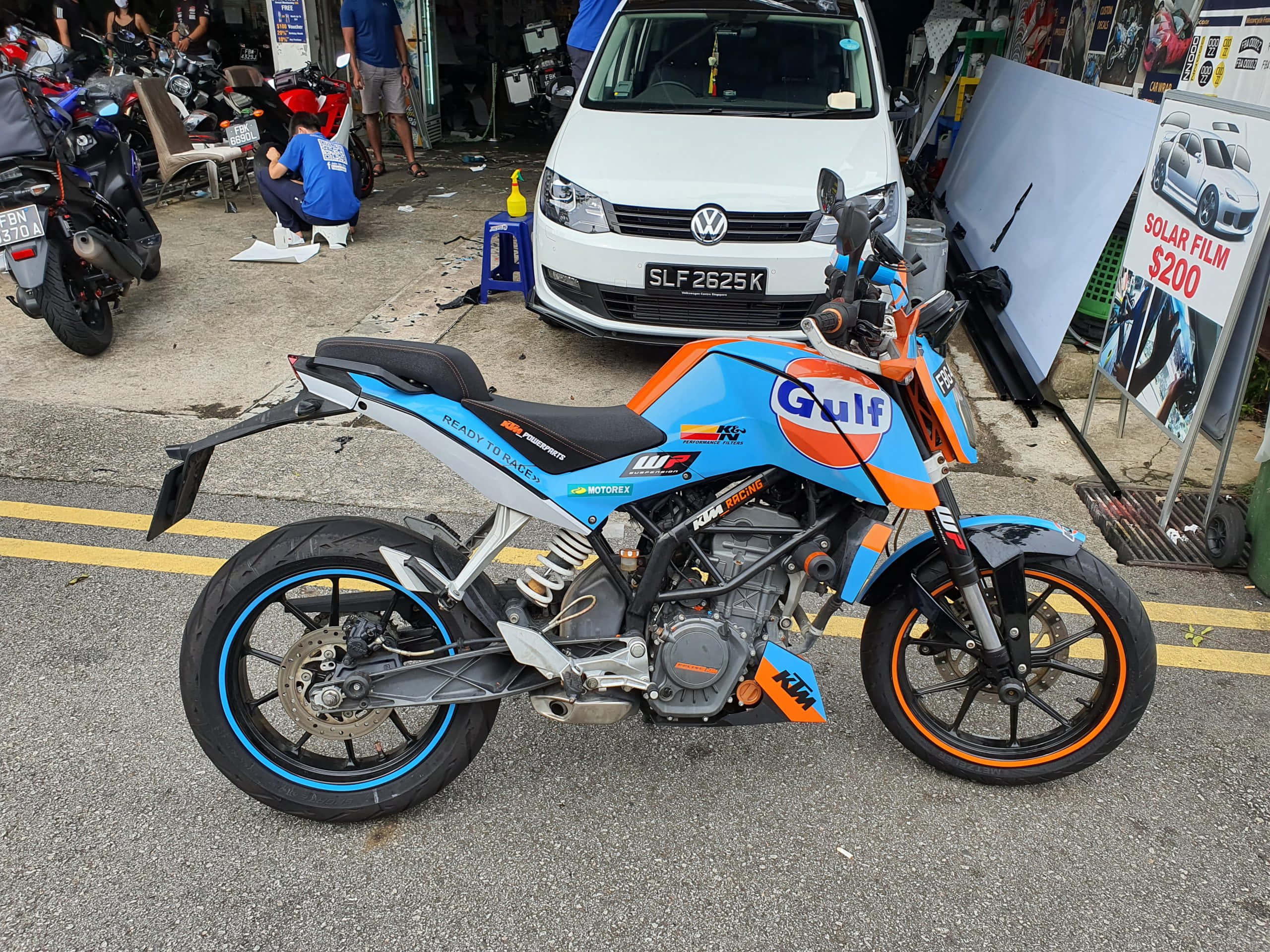 機車 桌布 壁紙 — 一輛帶有Gulf塗裝的鮮豔藍色和橙色KTM機車停放在鋪砌的停車場上，旁邊有一輛白色廂型車和其他機車。