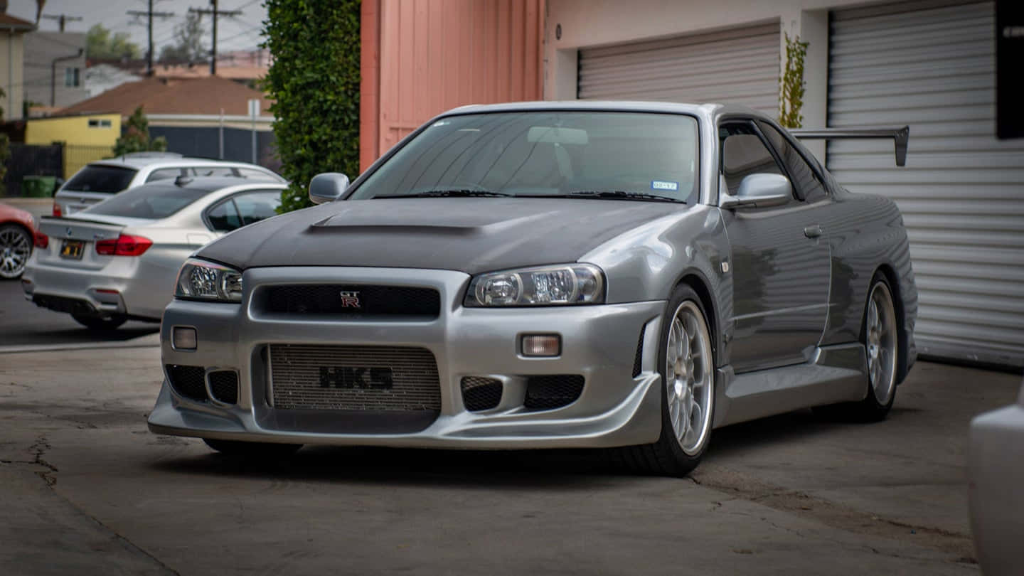 Paul Walker Skyline 桌布 壁紙 — 一輛帶有HKS標誌的時尚銀色日產Skyline R34停放在車庫旁的混凝土車道上。