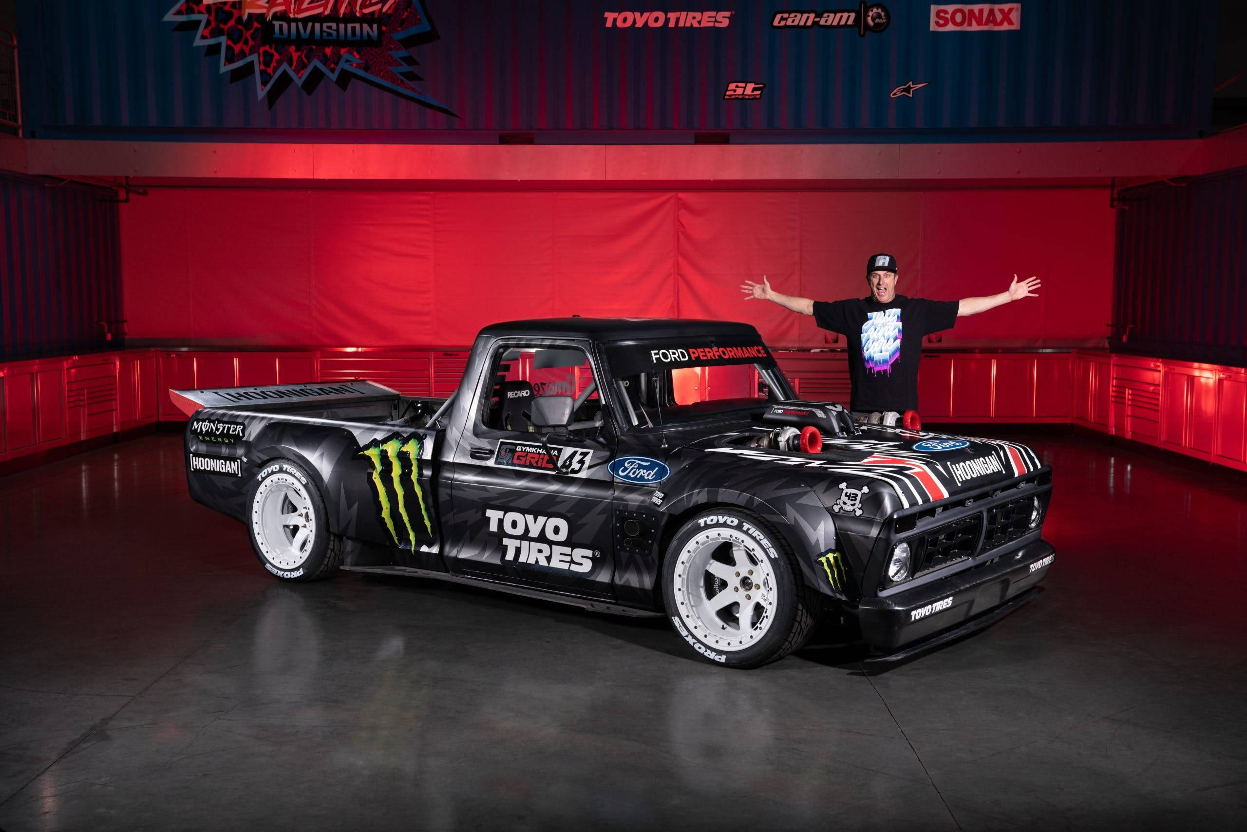 drifting 桌布 壁紙 — 一輛帶有Monster Energy和Toyo Tires貼紙的黑色漂移卡車在紅色燈光的車庫中展示，一名身穿黑色襯衫的男子勝利地站在它後面。