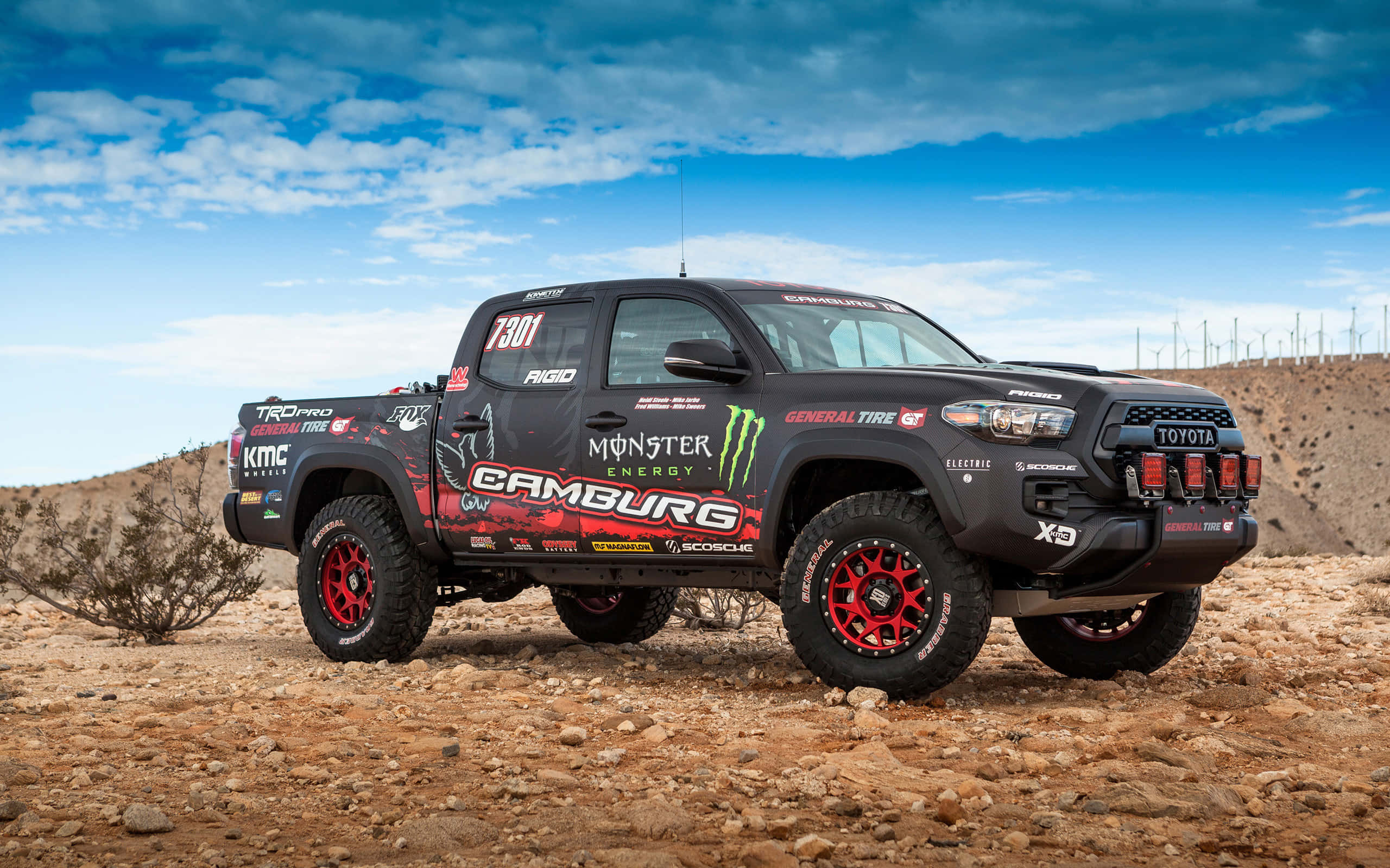 Toyota 桌布 壁紙 — 一輛帶有Monster Energy品牌標誌的黑色和紅色越野豐田Tacoma賽車卡車，在晴朗藍天和零星白雲下，屹立於岩石沙漠地形上。