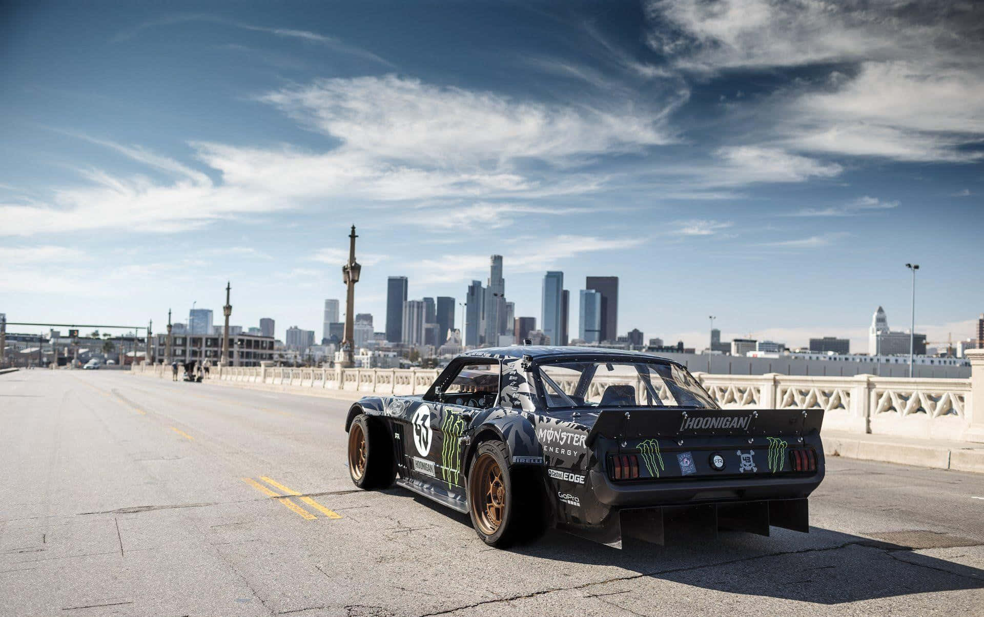 Hoonigan 桌布 壁紙 — 一輛帶有Monster Energy貼紙的黑色漂移車停在一座橋上，俯瞰著部分多雲藍天下的城市天際線。