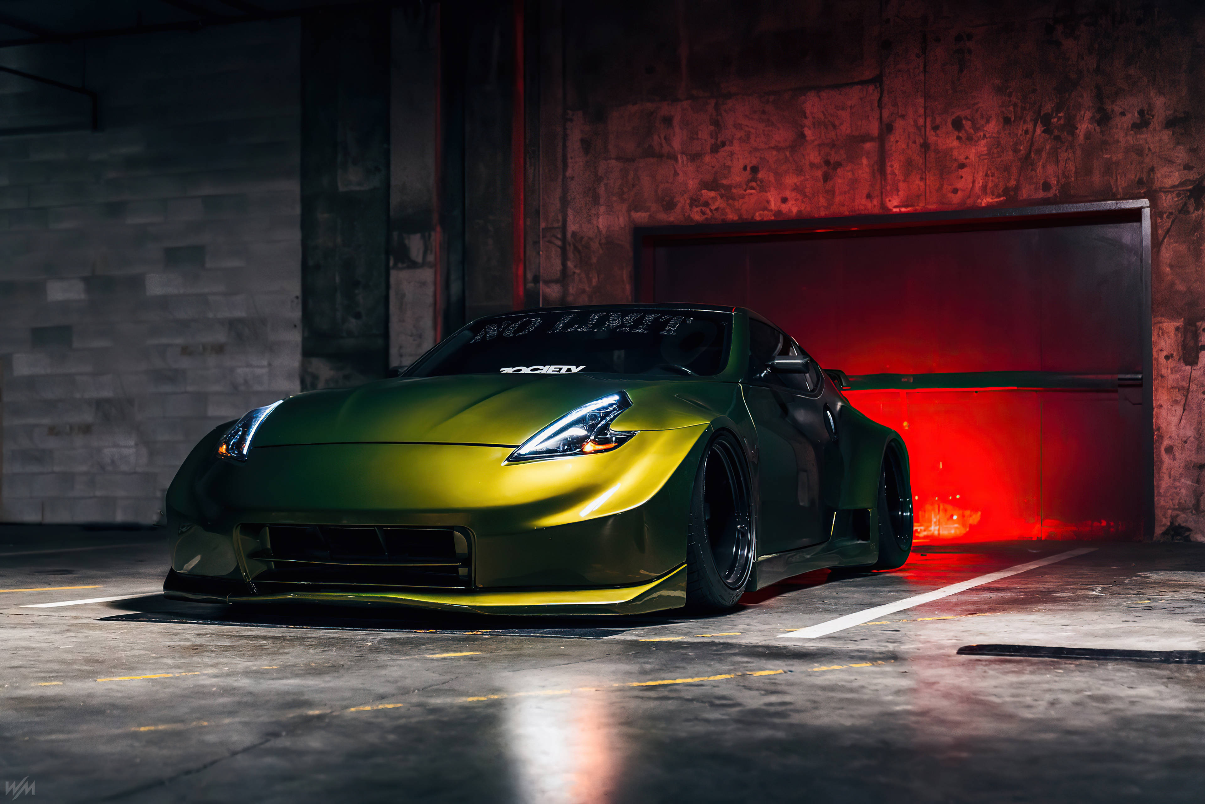 Nissan 370z 桌布 壁紙 — 一輛帶有定制寬體套件的啞光綠色跑車，戲劇性地停在昏暗的車庫中，沐浴在紅光下。