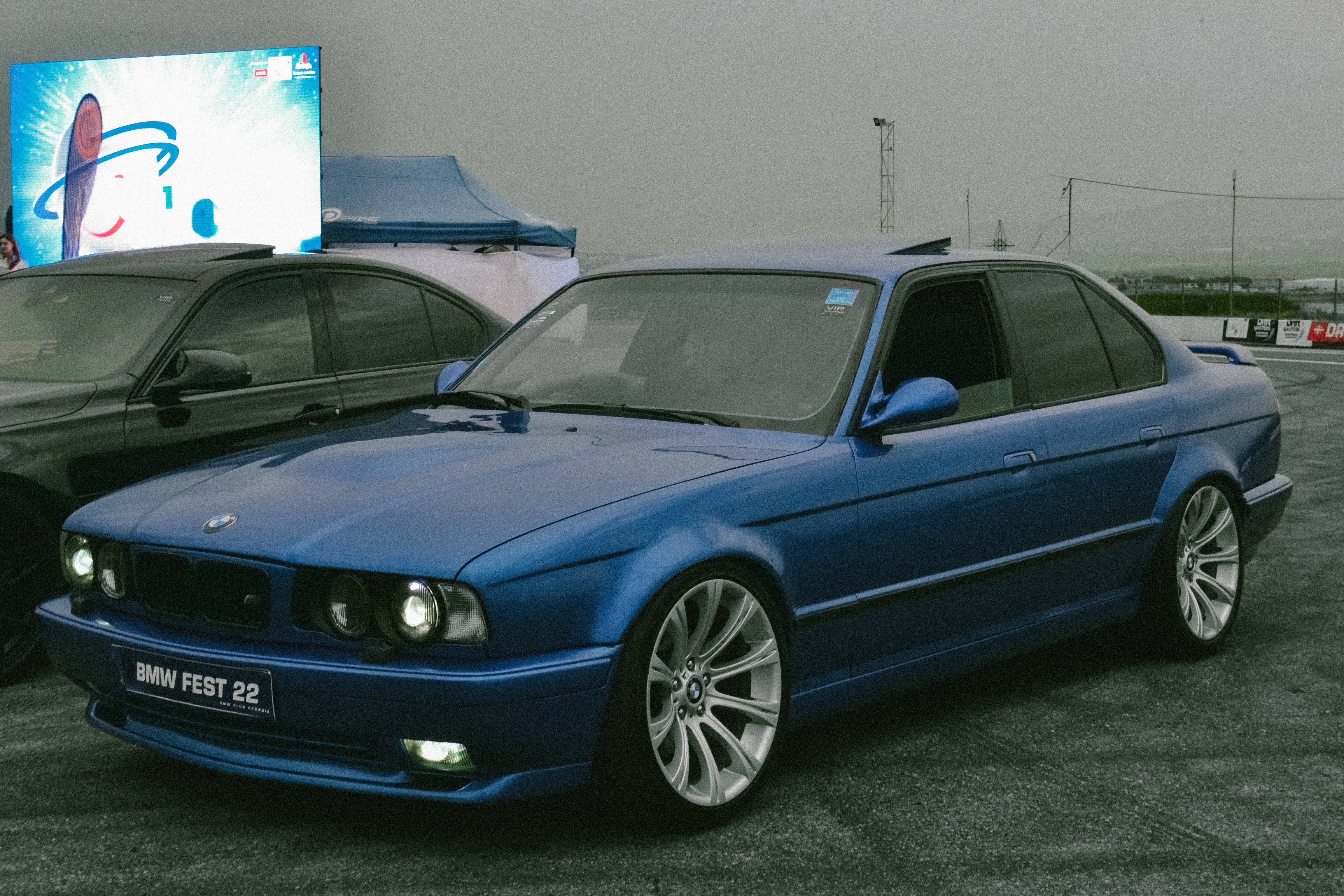 Racing 桌布 壁紙 — 一輛帶有定制輪圈的亮藍色BMW E36轎車，在多雲的天空下於汽車節上閃閃發光。