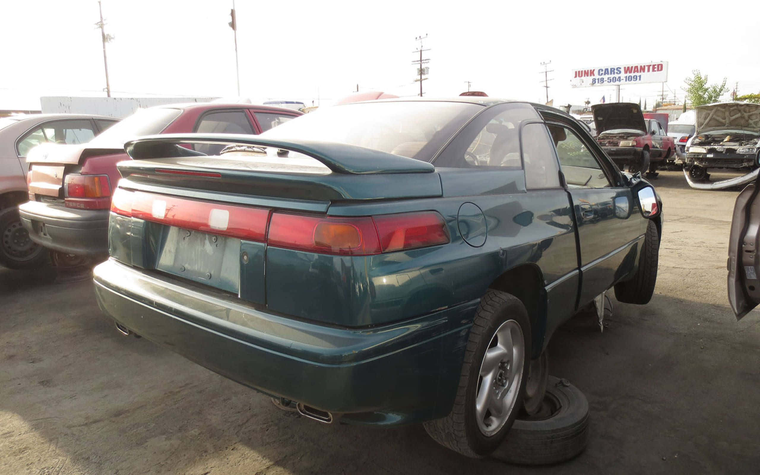 Subaru Svx 桌布 壁紙 — 一輛帶有後擾流板的青綠色運動型轎跑車被遺棄在廢車場，周圍是其他損毀的汽車，在明亮的日光下。