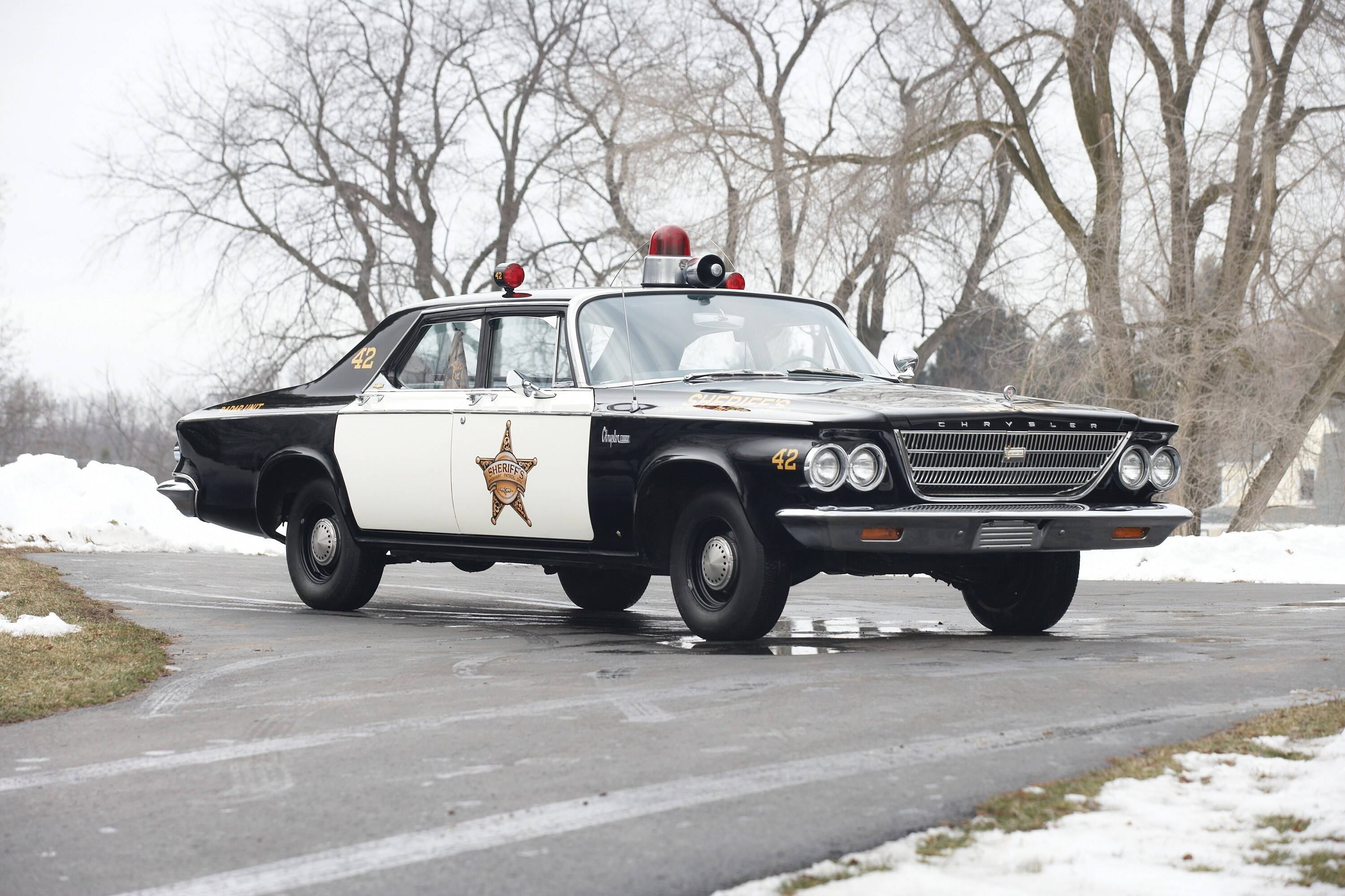 克萊斯勒 桌布 壁紙 — 一輛帶有星形徽章的復古黑白警長車在積雪的道路上行駛，道路兩旁是光禿禿的樹木。