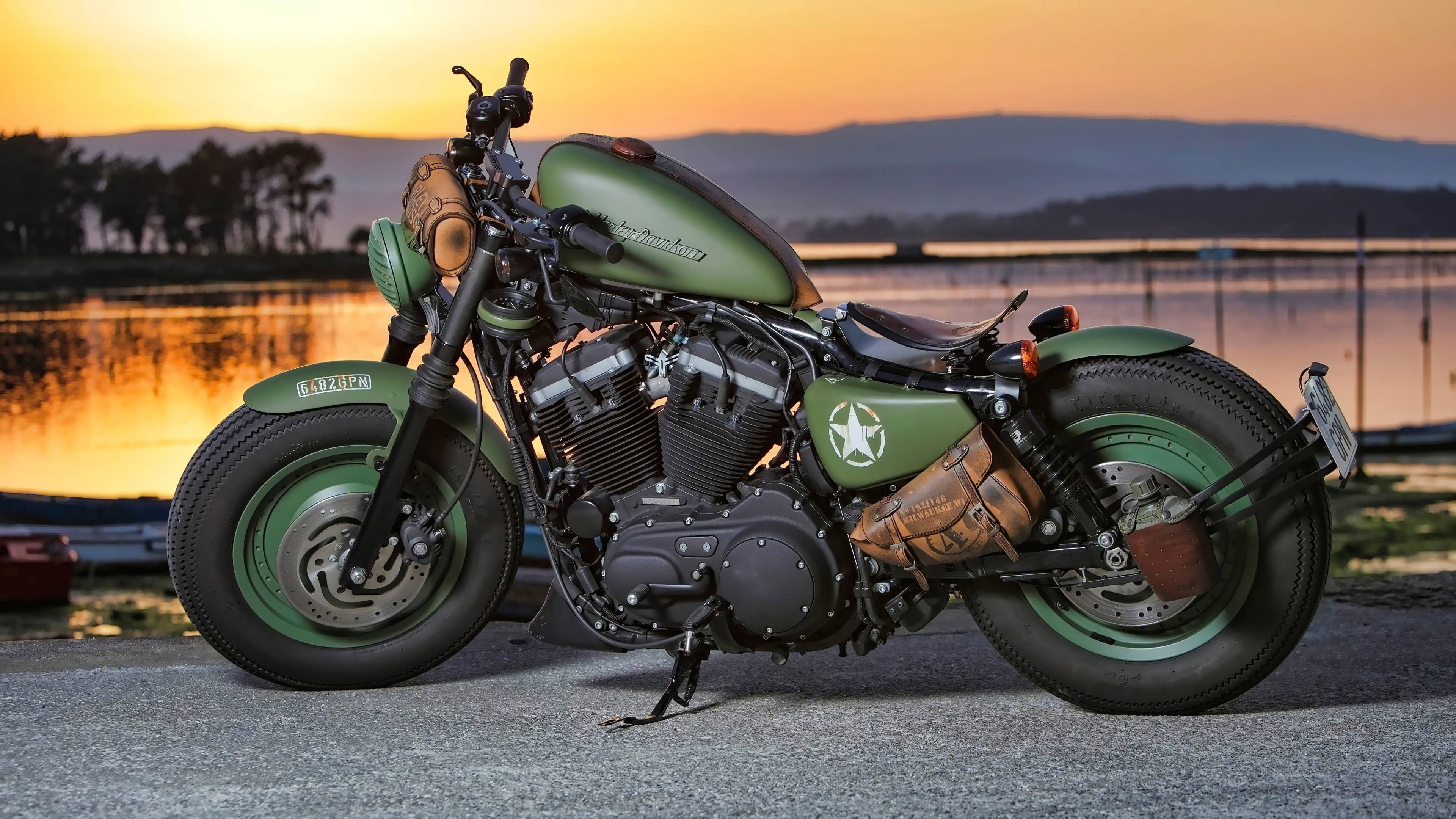 Harley 桌布 壁紙 — 一輛帶有星星標誌的啞光橄欖綠訂製摩托車，停在日落時分的湖邊，天空橙色，遠處有山丘。