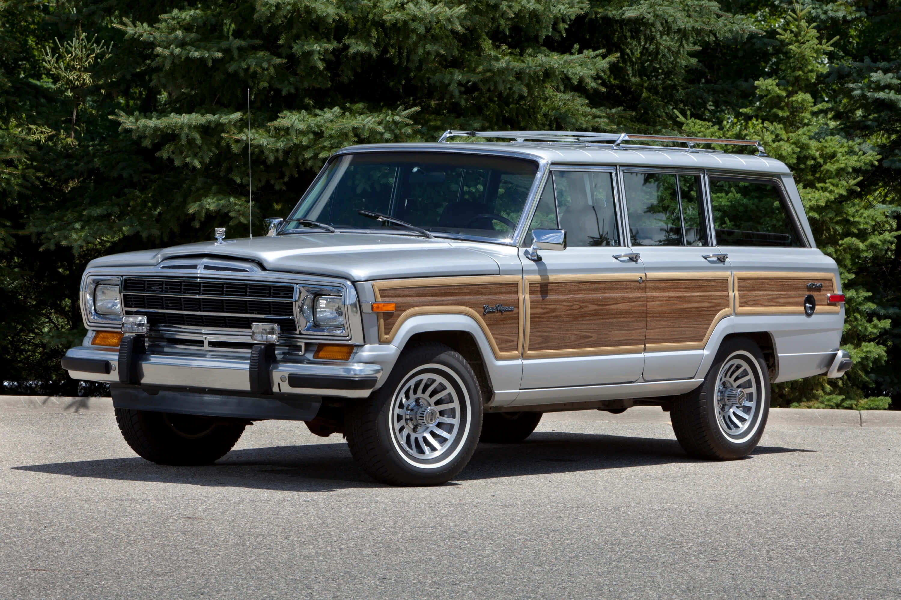 Jeep Wagoneer 桌布 壁紙 — 一輛帶有木飾板的銀色Jeep Wagoneer停泊在柏油路上，周圍是茂密的綠樹。