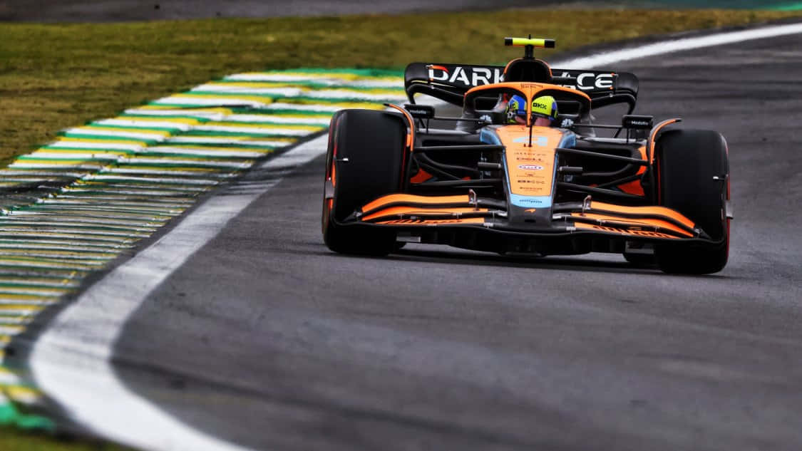 F1 桌布 壁紙 — 一輛帶有橙色和黑色塗裝的麥拉倫F1賽車在彎曲的瀝青賽道上奔馳，賽道兩旁有綠色和黃色的路緣石。