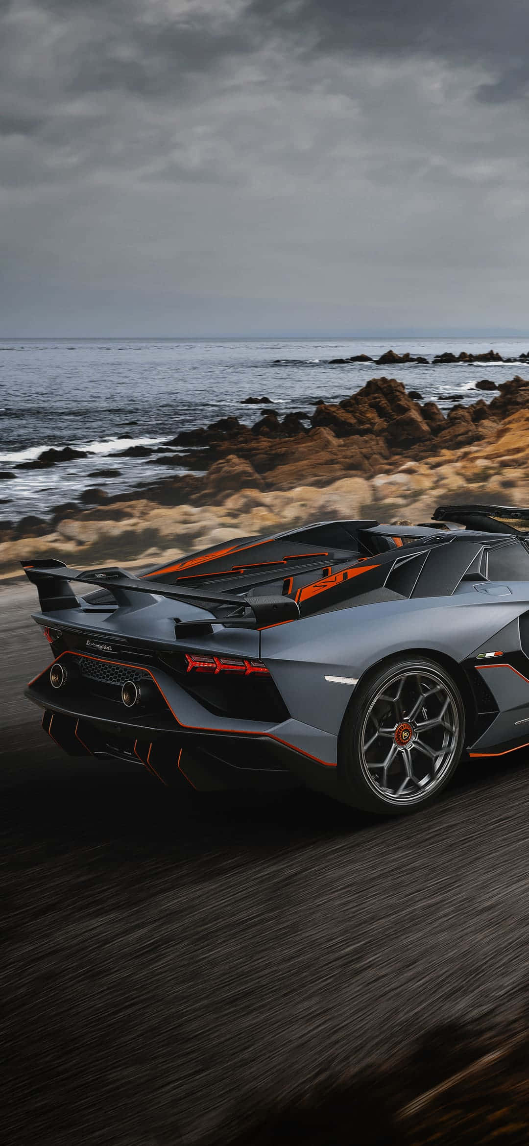 Pixel 3xl Lamborghini 桌布 壁紙 — 一輛帶有橙色點綴的時尚灰色藍寶堅尼在暴風雨的天空下沿著海岸公路飛馳。