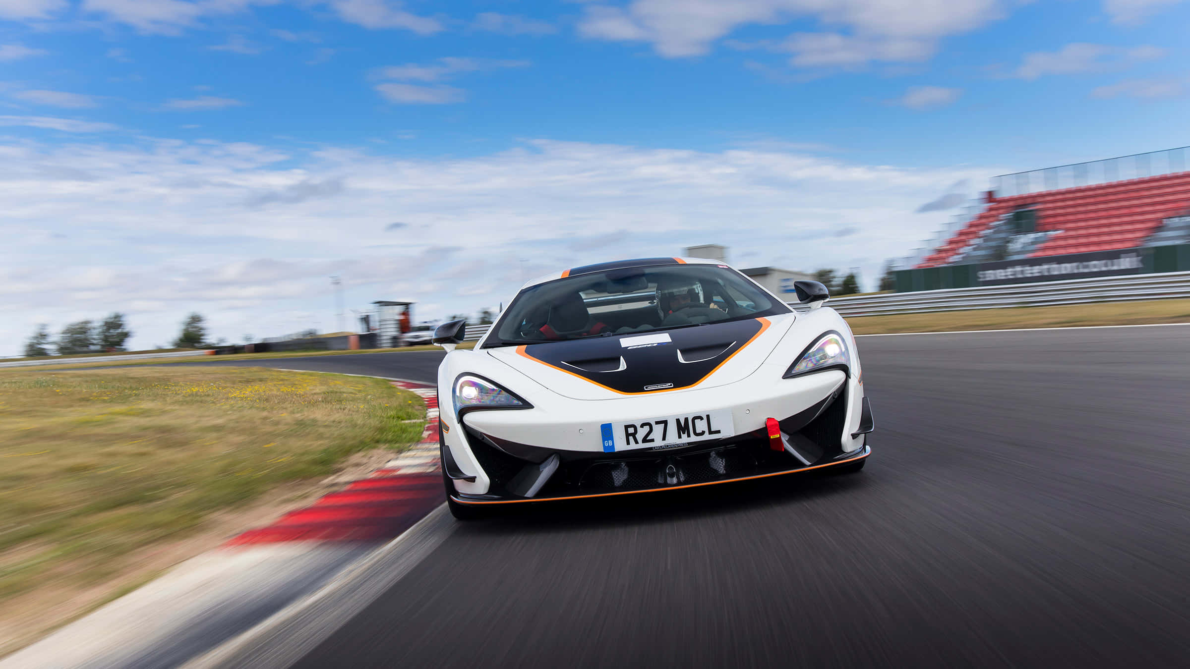 Mclaren 620r 桌布 壁紙 — 一輛帶有橙色點綴的白色McLaren跑車在明亮的藍天下，以動態模糊效果在賽道上奔馳。