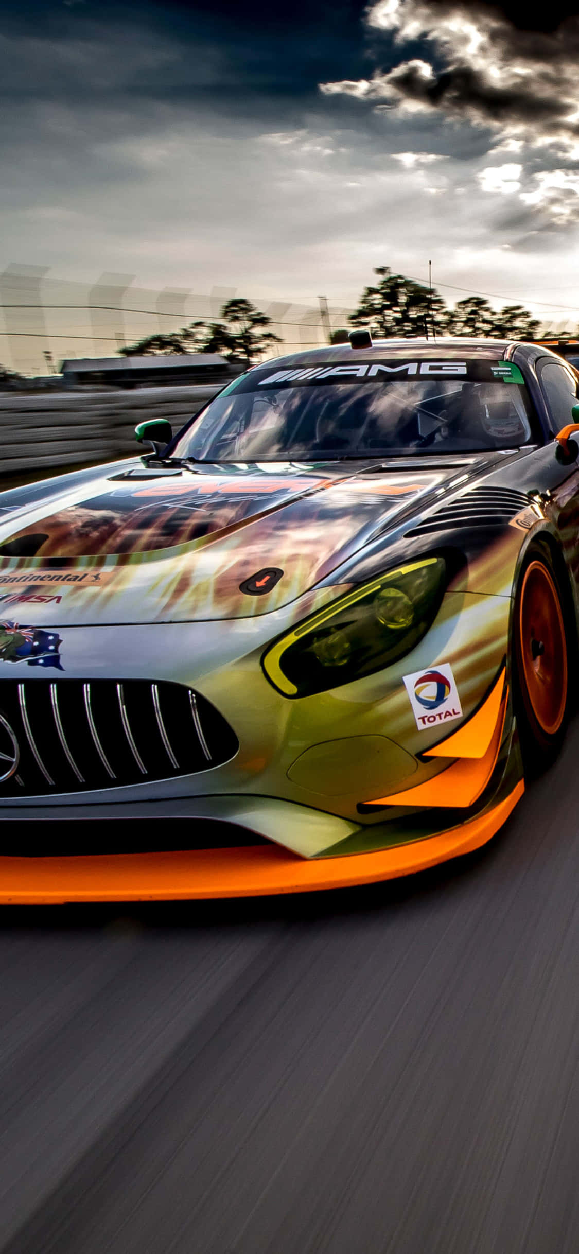 iPhone X Amg Gt-r 桌布 壁紙 — 一輛帶有火熱橙色和金色塗裝的Mercedes-AMG GT3賽車，在戲劇性的多雲天空下沿賽道飛馳。