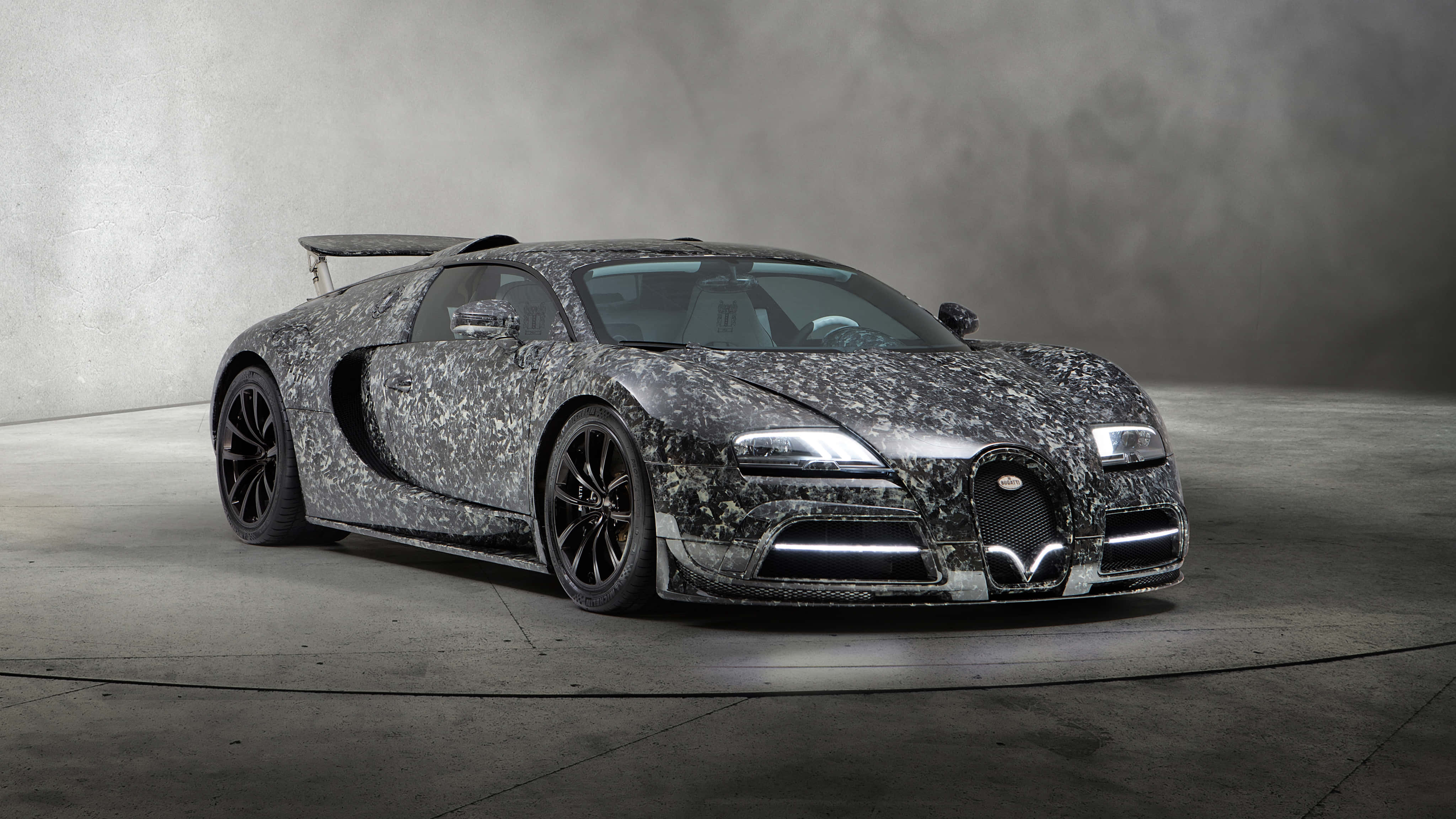 Best Bugatti 桌布 壁紙 — 一輛帶有灰色和黑色斑點迷彩圖案的Bugatti超級跑車，在極簡主義的混凝土工作室中，於戲劇性的燈光下靜止不動。