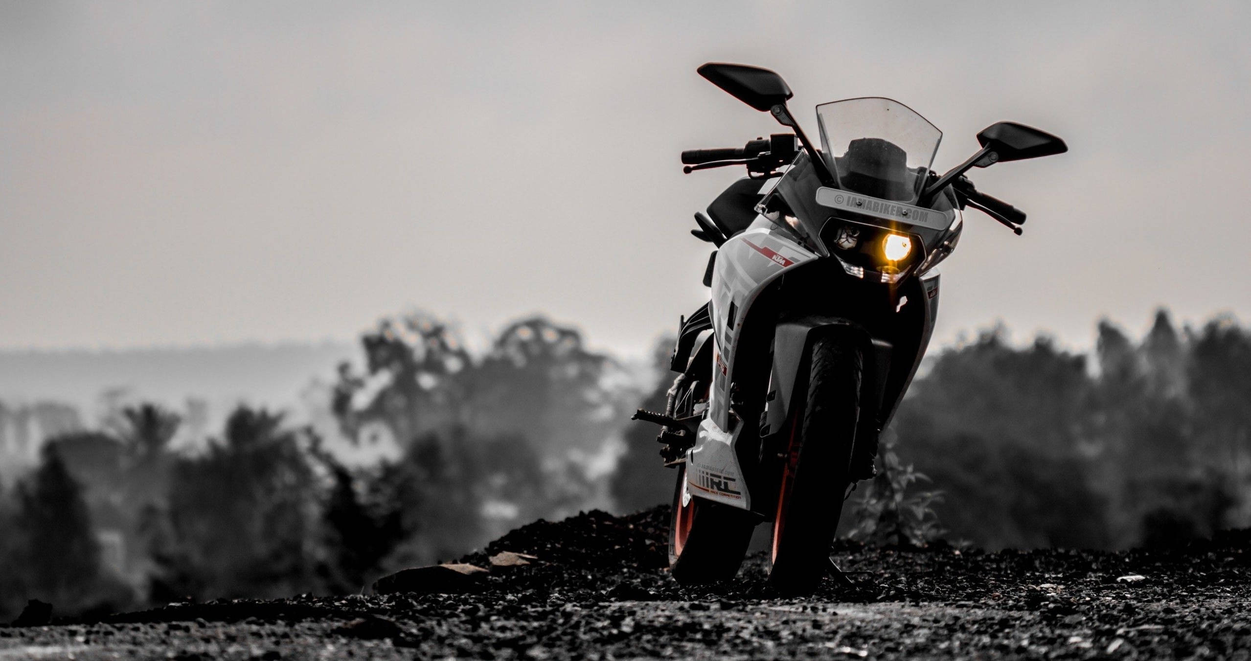 Ktm Bike 桌布 壁紙 — 一輛帶有發光頭燈的運動機車，佇立在碎石山脊上，背景是朦朧的單色風景。