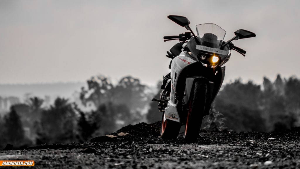 Ktm Rc 390 桌布 壁紙 — 一輛帶有發光頭燈的運動機車，佇立在碎石山丘上，背景是朦朧的黑白風景。