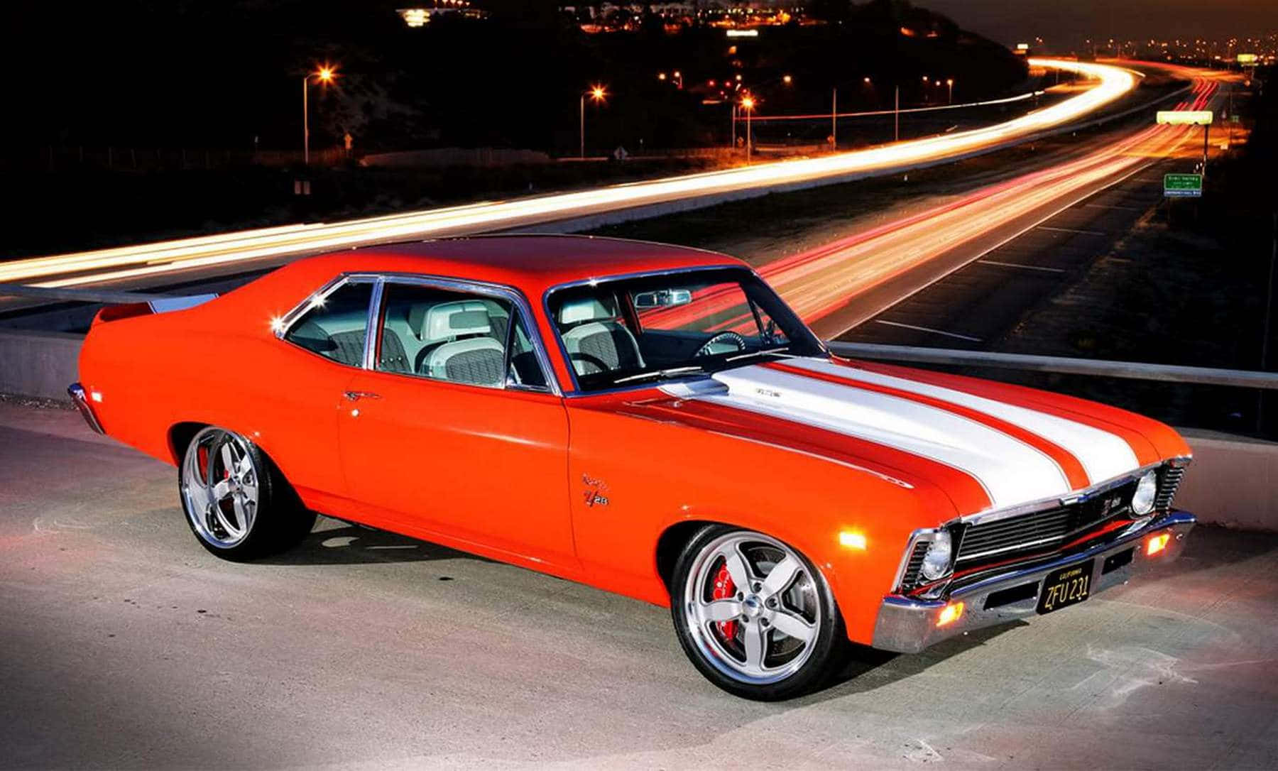 chevy 桌布 壁紙 — 一輛帶有白色條紋的鮮豔橙色經典肌肉車停在夜間高速公路上，伴隨著移動車輛的光軌。