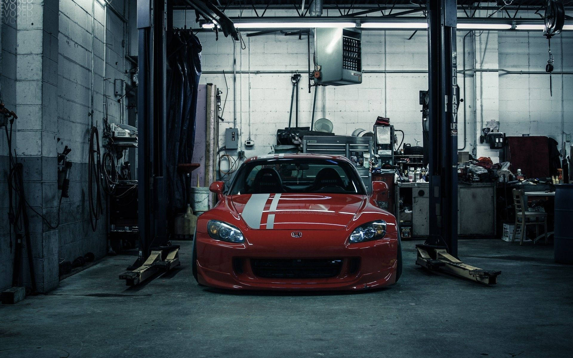 JDM 桌布 壁紙 — 一輛帶有白色賽車條紋的紅色跑車位於昏暗、雜亂的汽車修理廠的升降機中央。