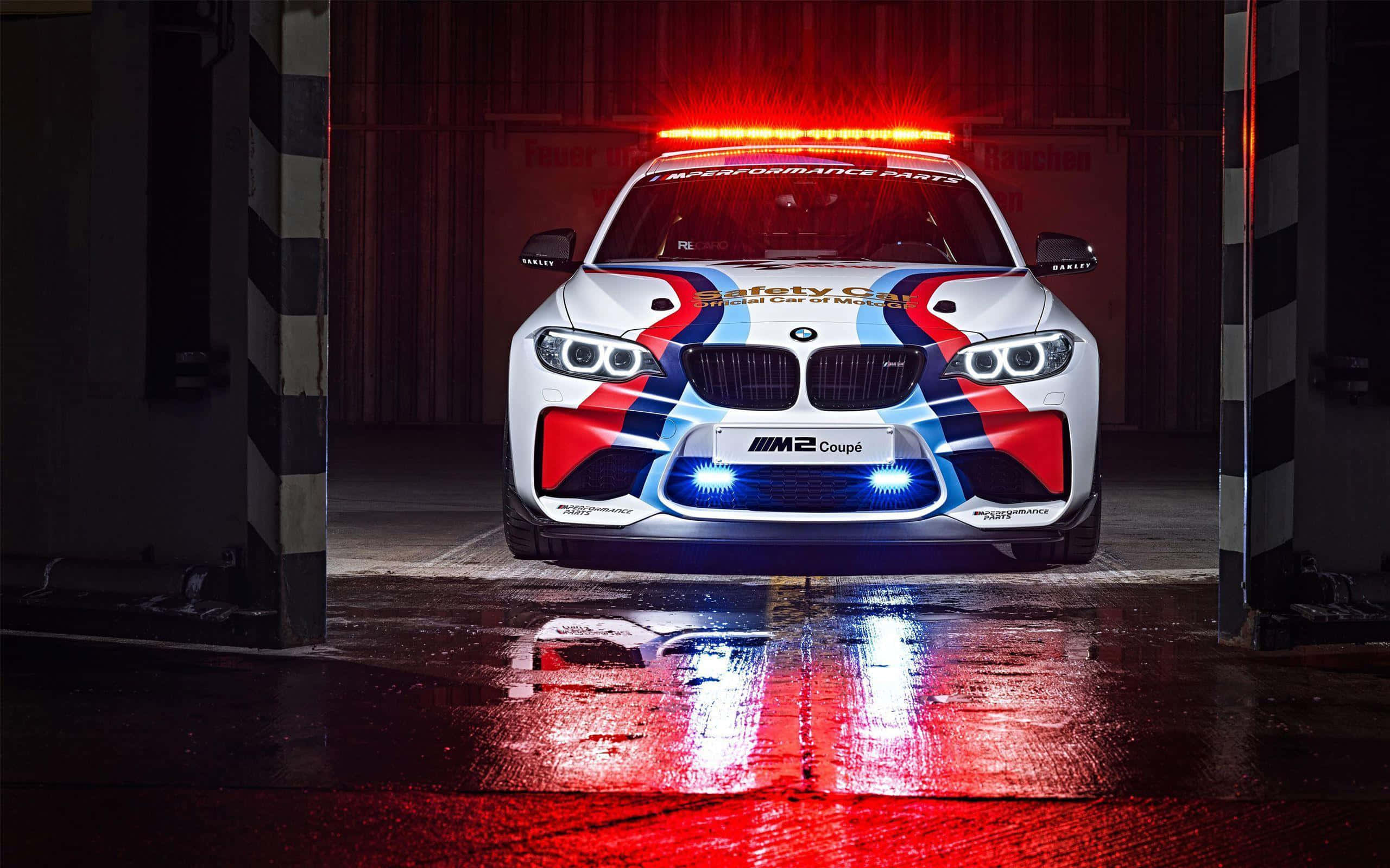Safety Car 桌布 壁紙 — 一輛帶有紅、藍、黑色塗裝的白色BMW M2 Coupe安全車，停在潮濕的車庫中，被發出紅光和藍光的燈光照亮，光線反射在地板上。
