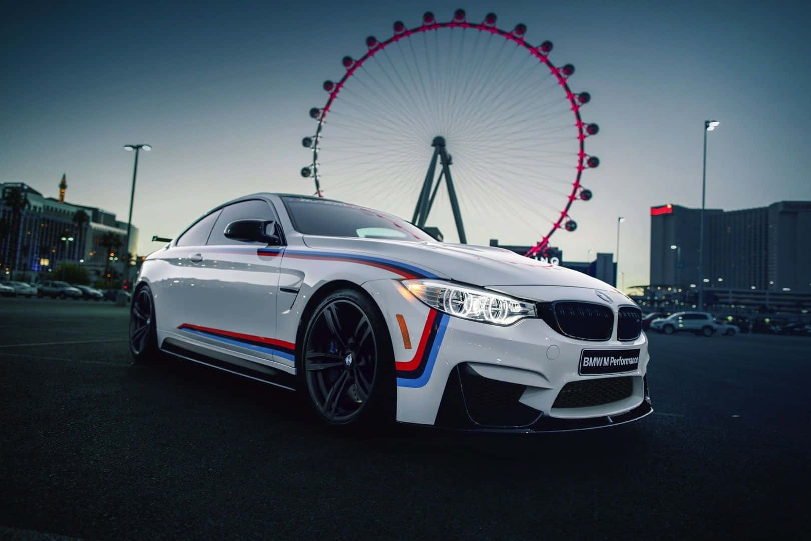 BMW 動圖 桌布 壁紙 — 一輛帶有紅色、藍色和橙色條紋的白色BMW M4 Performance雙門跑車，在黃昏時停泊在拉斯維加斯High Roller摩天輪前。