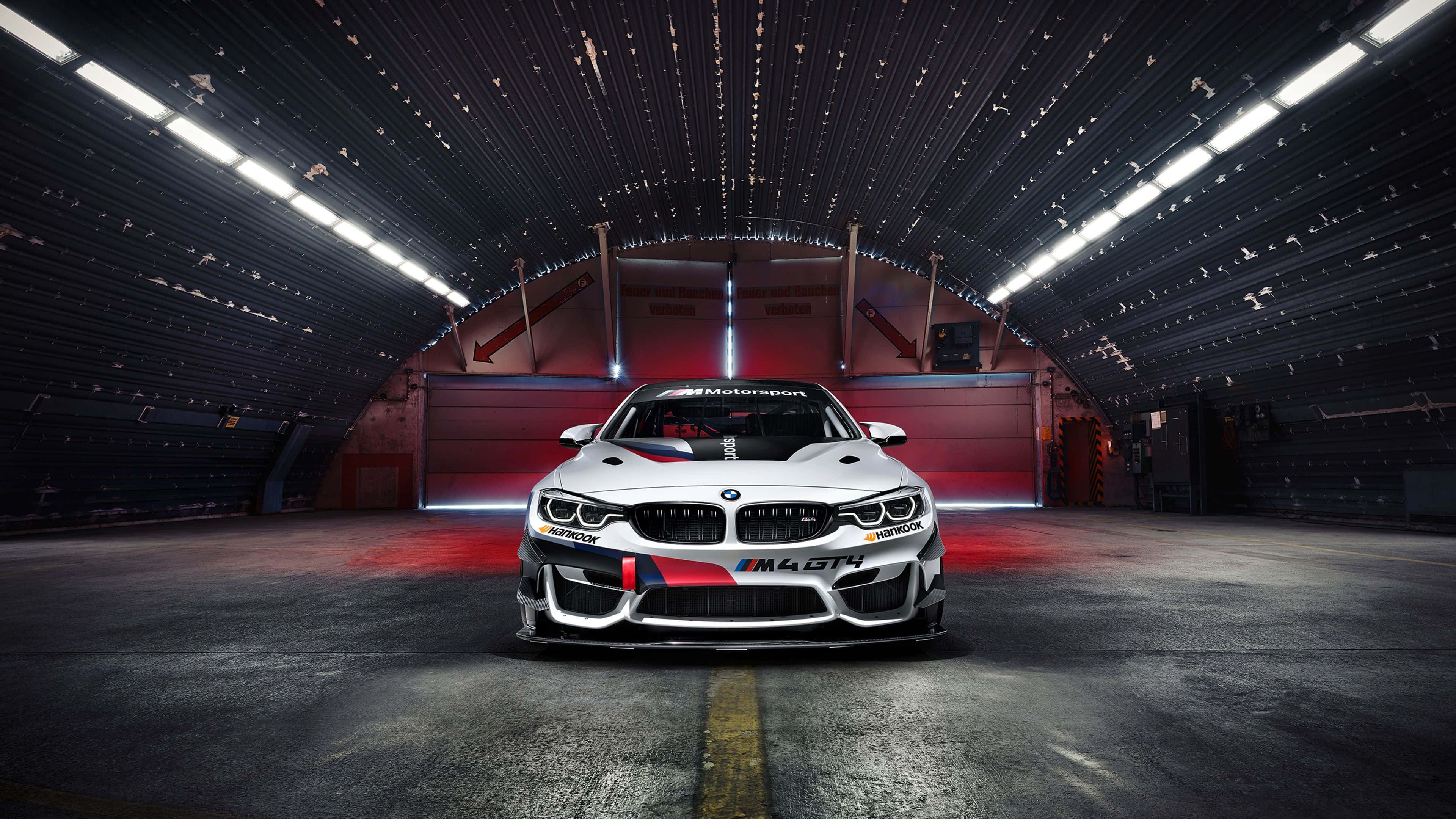 4K BMW 壁紙 桌布 壁紙 — 一輛帶有紅色和藍色點綴的白色BMW M4 GT4賽車，在明亮的頂燈下位於黑暗的工業車庫中央。