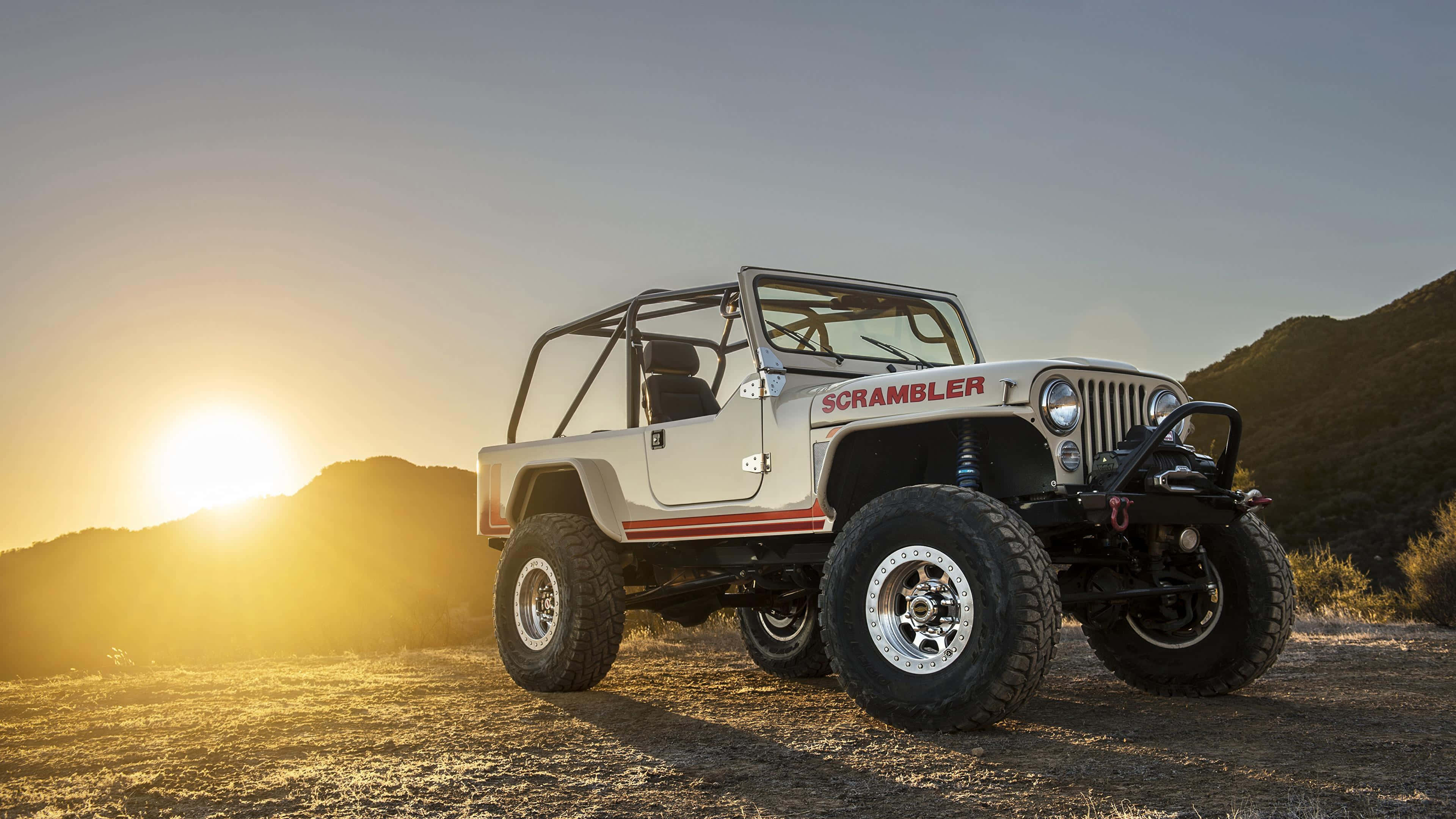 Jeep Cj 桌布 壁紙 — 一輛帶有紅色條紋的白色Jeep Scrambler停在塵土飛揚的小徑上，夕陽在起伏的山丘後落下，灑下金色的光芒。