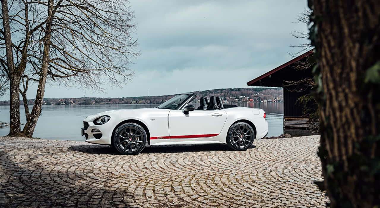 Fiat 124 Spider 桌布 壁紙 — 一輛帶有紅色條紋的白色敞篷跑車停泊在鵝卵石小徑上，旁邊是湖，背景有光禿禿的樹木和木屋。