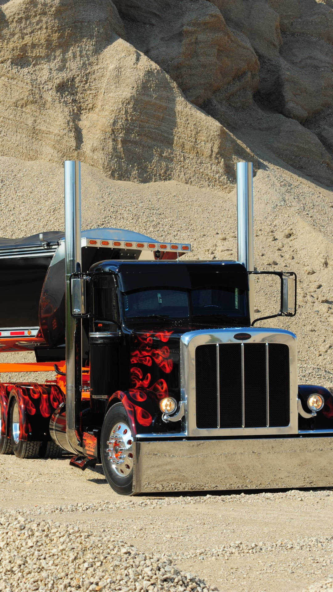 Peterbilt 桌布 壁紙 — 一輛帶有紅色火焰貼花的黑色半掛卡車停在礫石上，背景是陽光照耀的岩石峭壁。