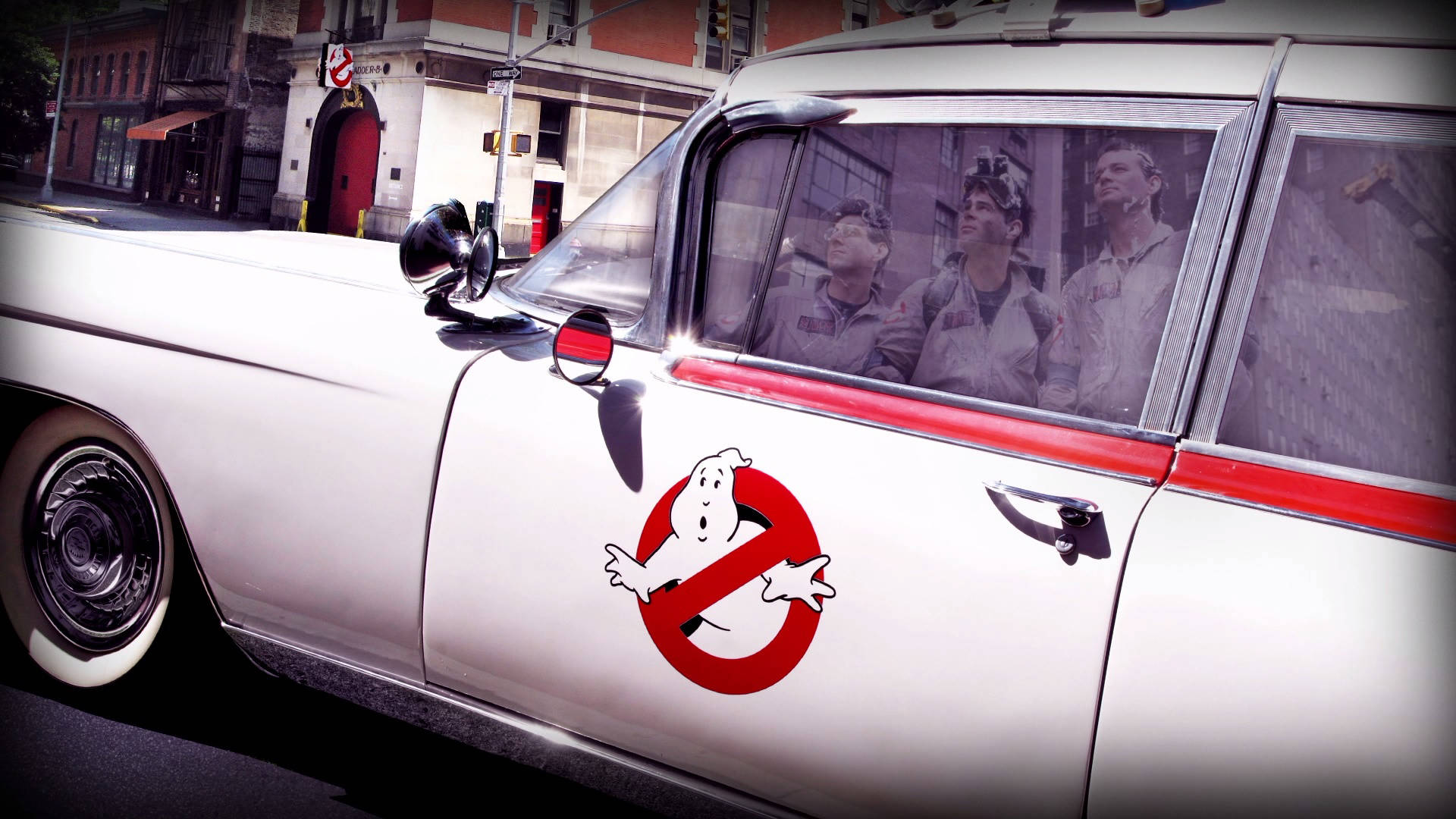 Ghostbusters 桌布 壁紙 — 一輛帶有紅色飾邊和Ghostbusters標誌的白色1950年代靈車行駛在城市的街道上，車內可見三名穿著連身工作服的男人。