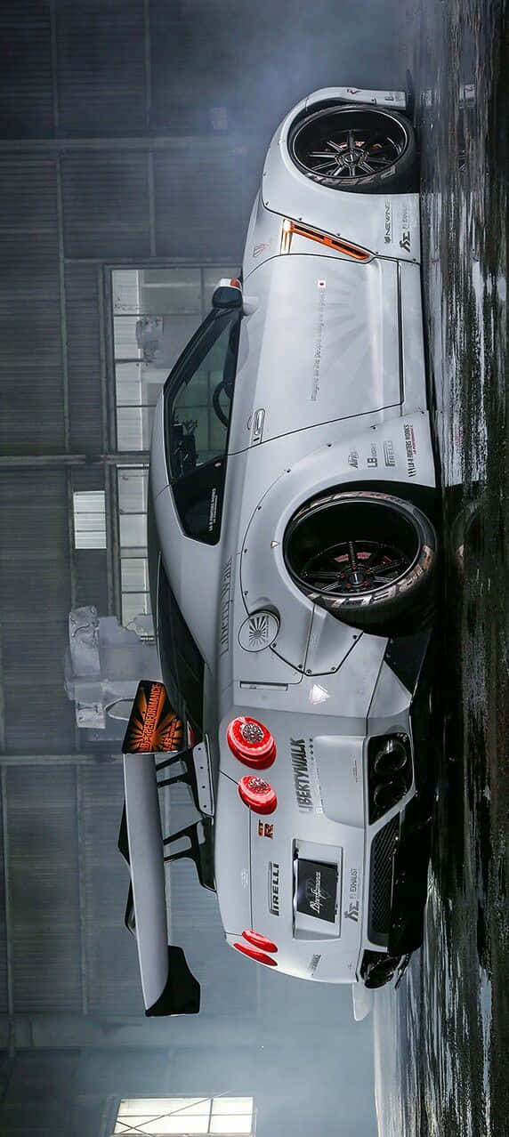 Nissan R35 Gtr 桌布 壁紙 — 一輛帶有紅色點綴和贊助商標誌的白色賽車停放在昏暗的工業車庫中，地面濕滑，蒸汽升騰。