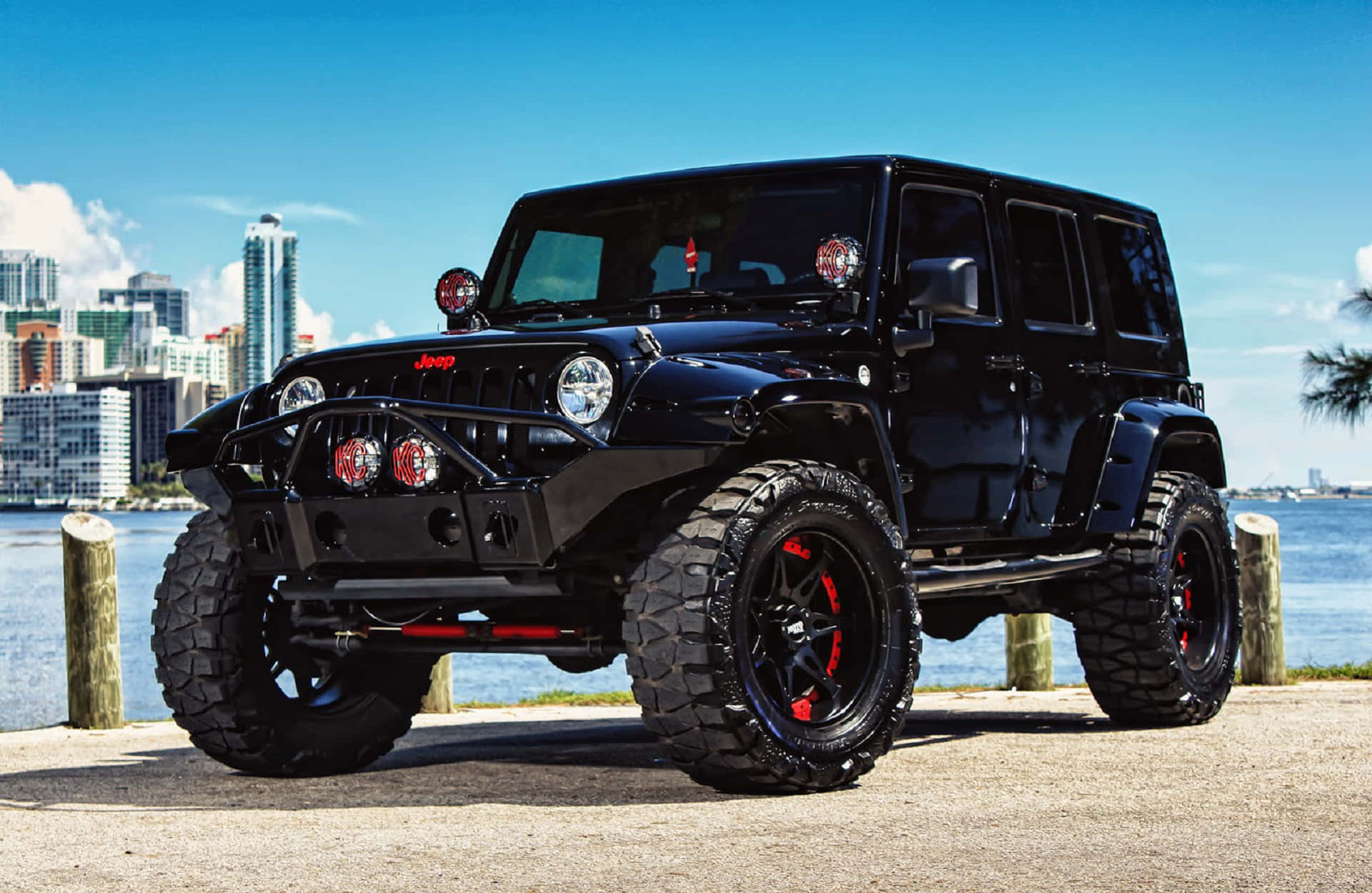 黑色吉普車 桌布 壁紙 — 一輛帶有紅色點綴和超大輪胎的黑色Jeep Wrangler停在濱海碼頭上，背景是晴朗藍天下的城市天際線。