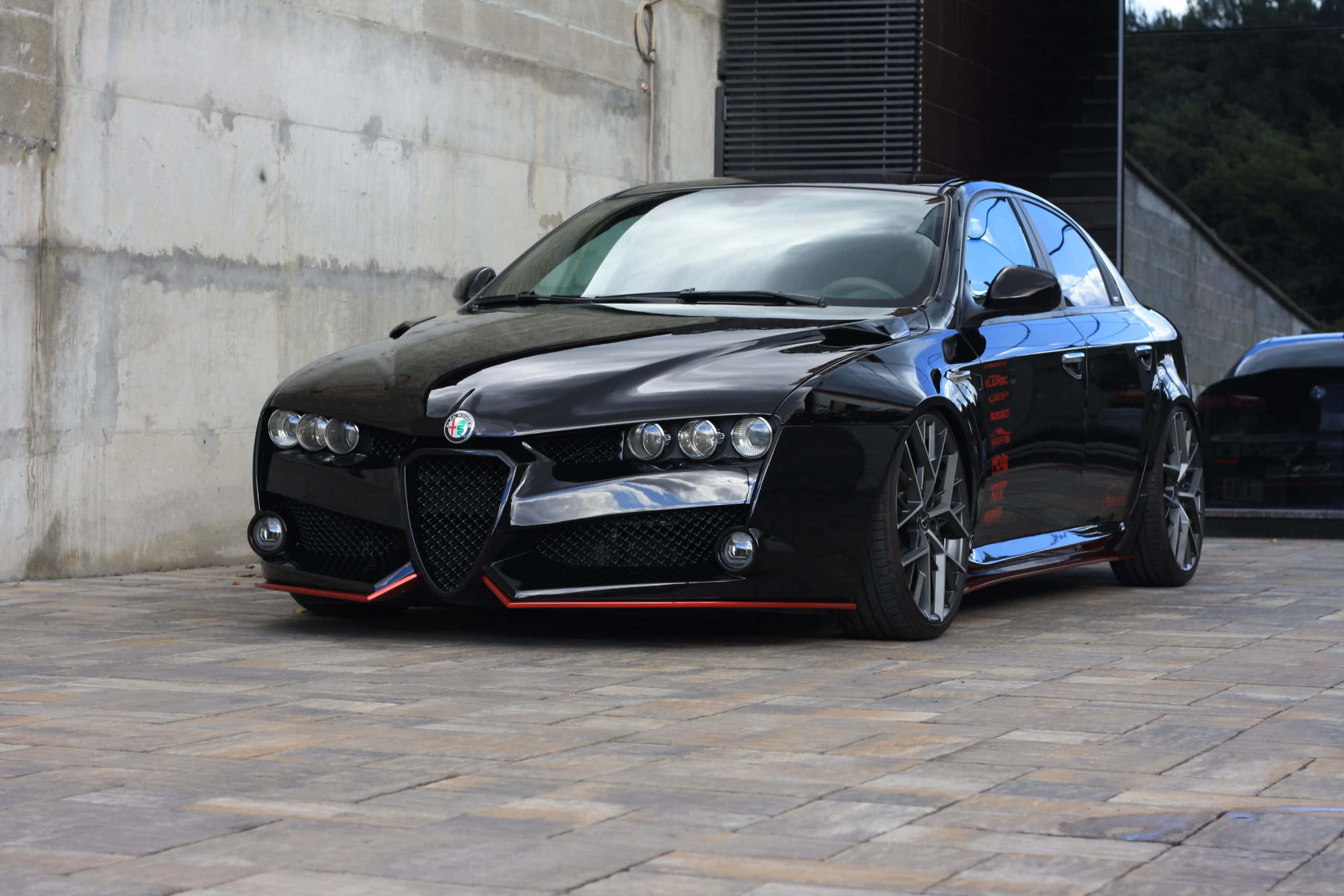 Alfa Romeo 159 桌布 壁紙 — 一輛帶有紅色點綴的亮黑色愛快羅密歐轎車停在混凝土牆旁的石板路上。