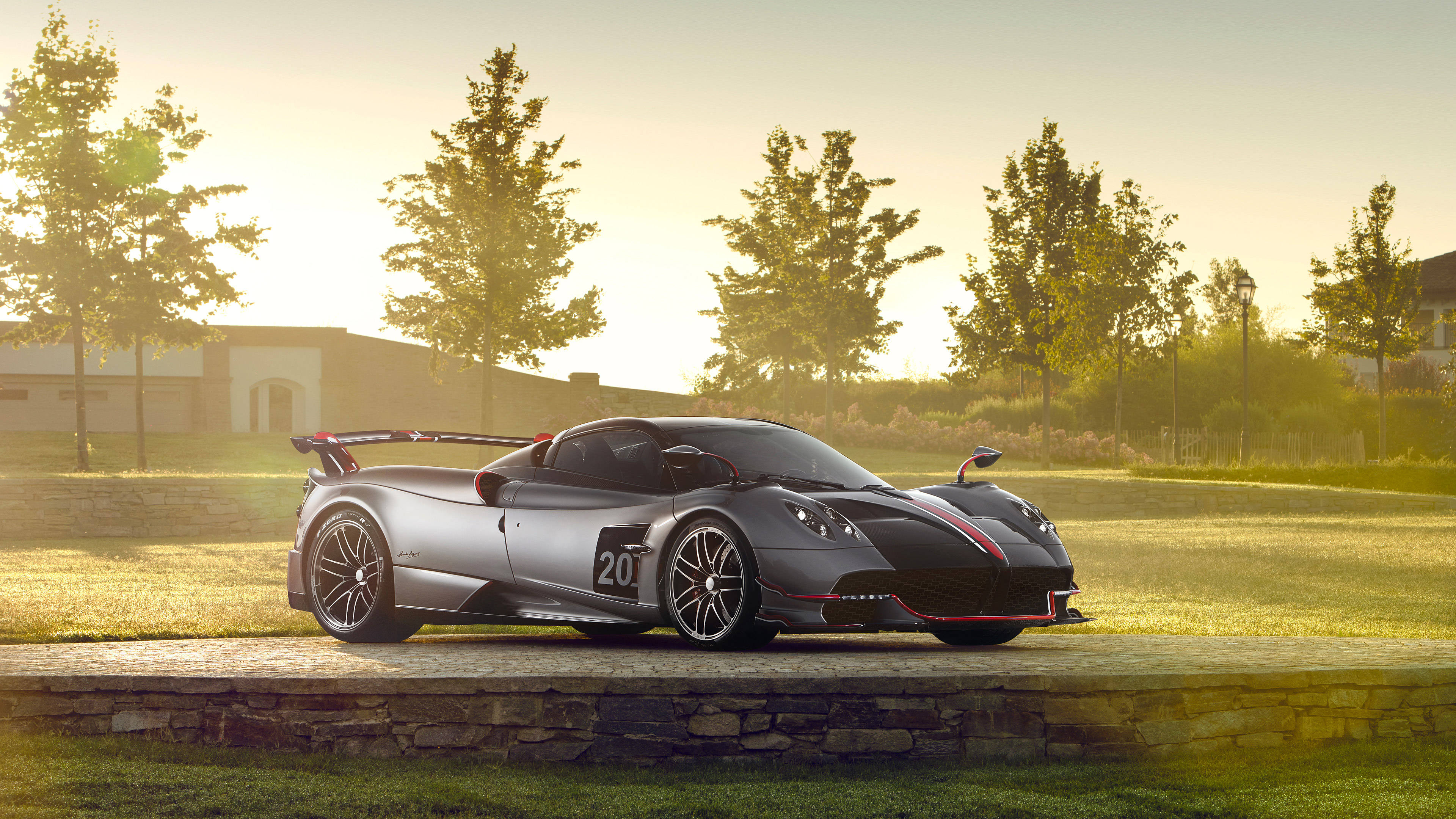 pagani iPhone 桌布 壁紙 — 一輛帶有紅色點綴的時尚銀色Pagani超跑在黃金時刻停放在石牆上，被樹木和柔和陽光環繞。