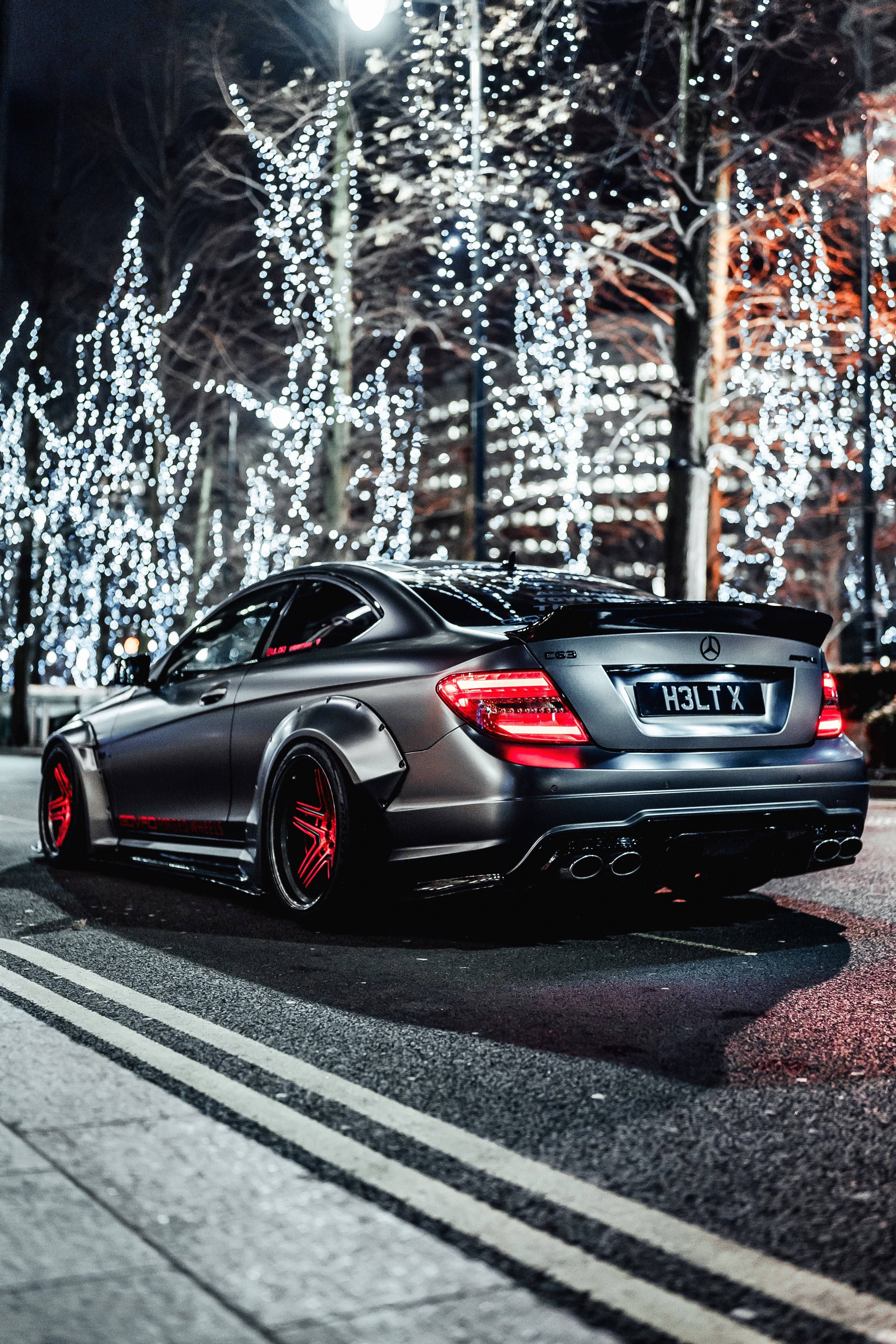 AMG iPhone 桌布 壁紙 — 一輛帶有紅色點綴的消光黑色梅賽德斯轎跑車，夜間停在積雪樹木下閃爍的串燈下。