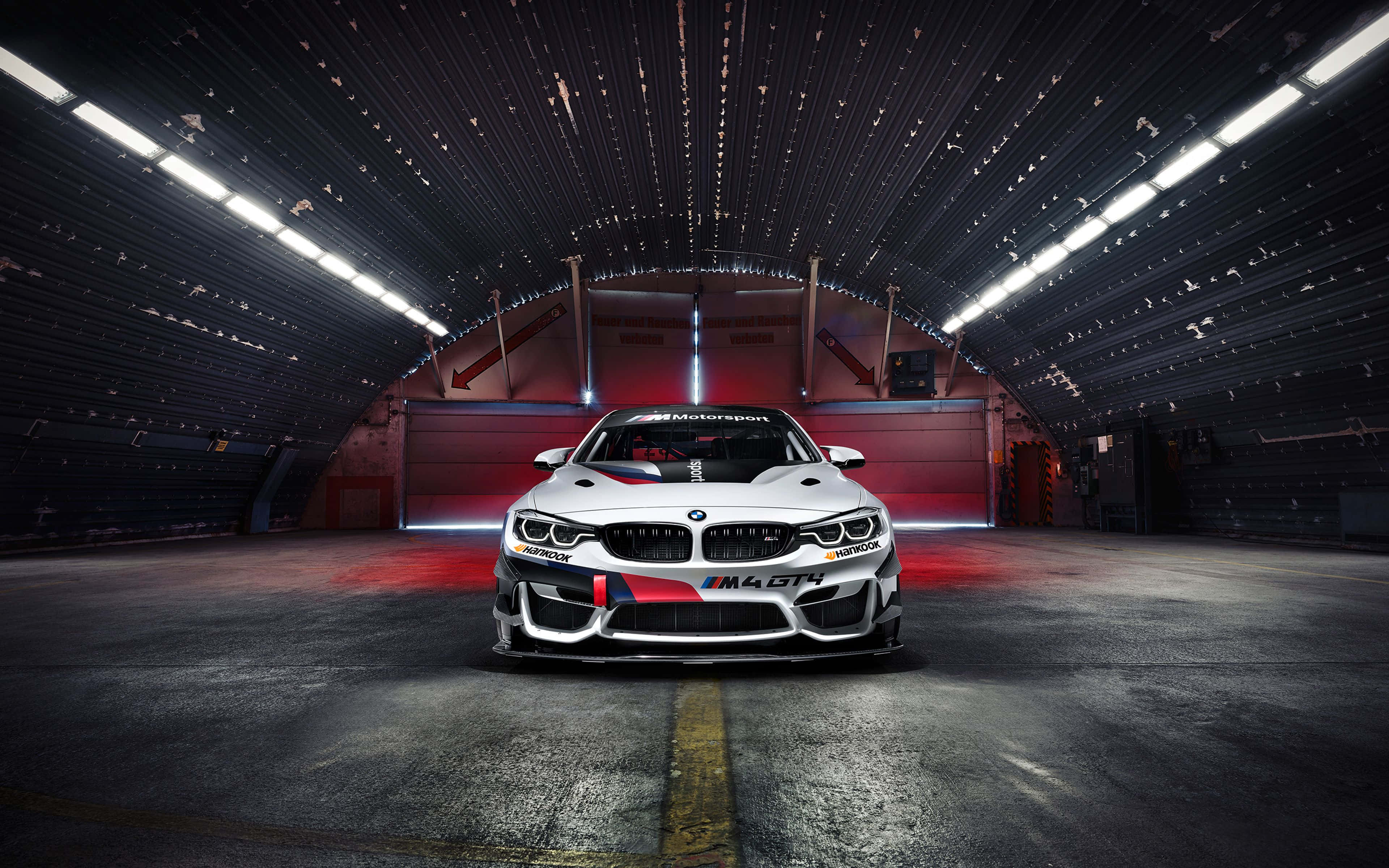 跑車 桌布 壁紙 — 一輛帶有紅色點綴的白色BMW M4 GT4賽車，在明亮的頂燈下居中停放在黑暗的工業車庫中。