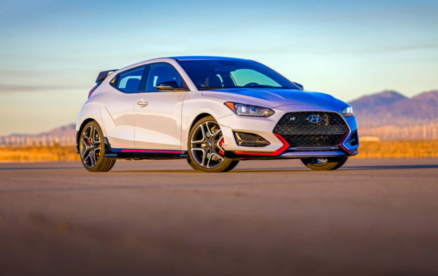 Hyundai 桌布 壁紙 — 一輛帶有紅色點綴的白色現代Veloster N跑車停在沙漠公路上，背景是夕陽和山脈。