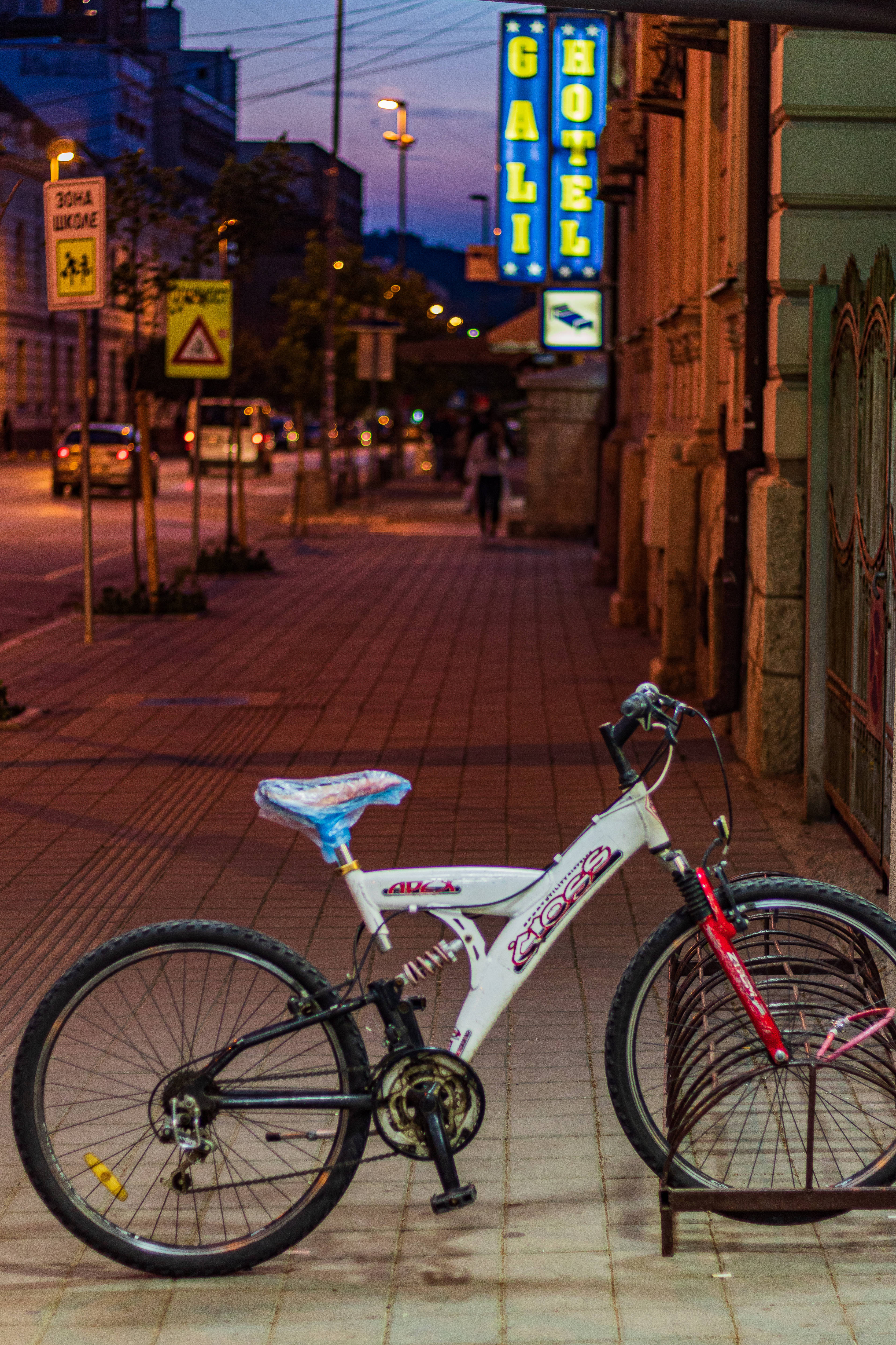 Serbia 桌布 壁紙 — 一輛帶有紅色點綴的白色自行車停放在黃昏時分寧靜的城市人行道上的自行車架上，被溫暖的路燈和發光的酒店招牌照亮。