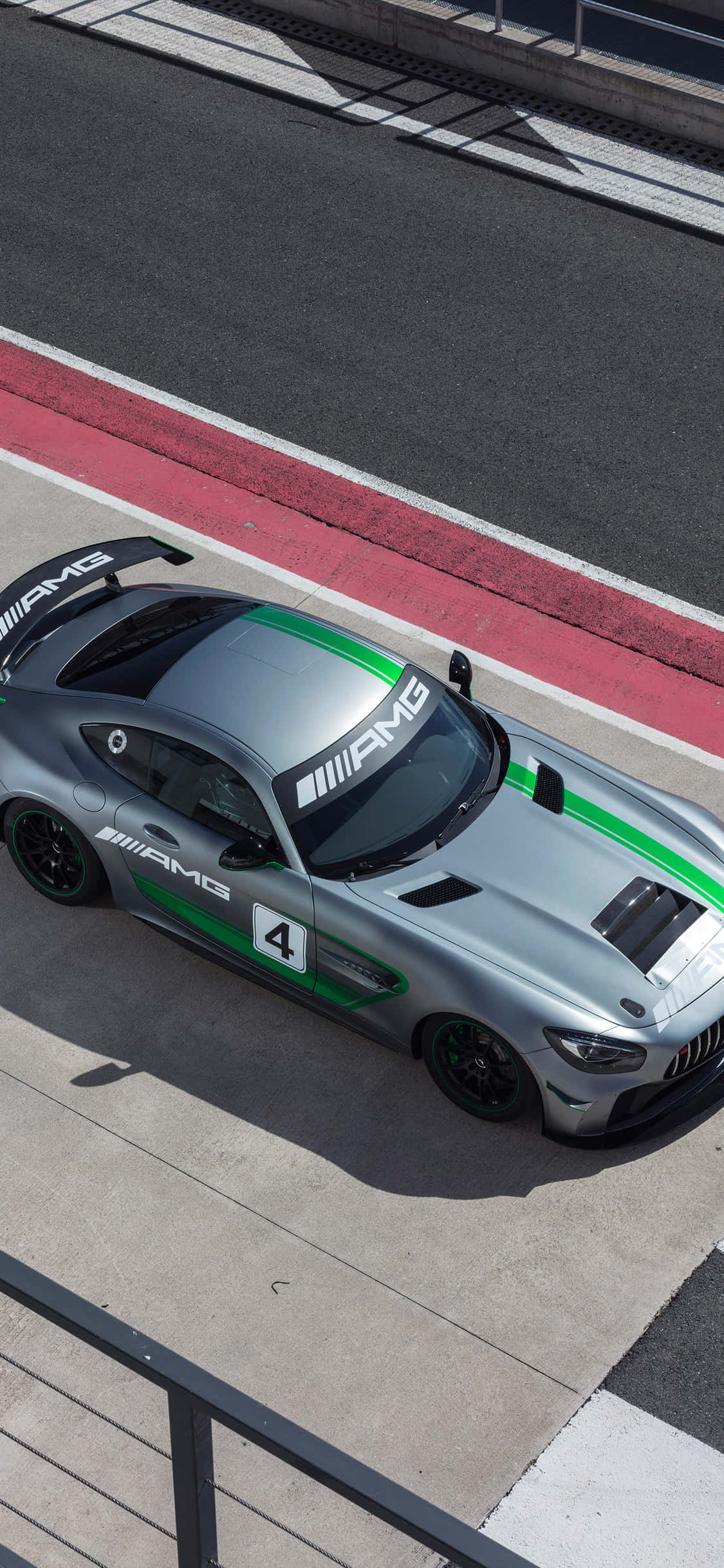 iPhone X Amg Gt-r 桌布 壁紙 — 一輛帶有綠色條紋的時尚銀色AMG賽車在明亮的日光下沿著紅色路緣的賽道飛馳。