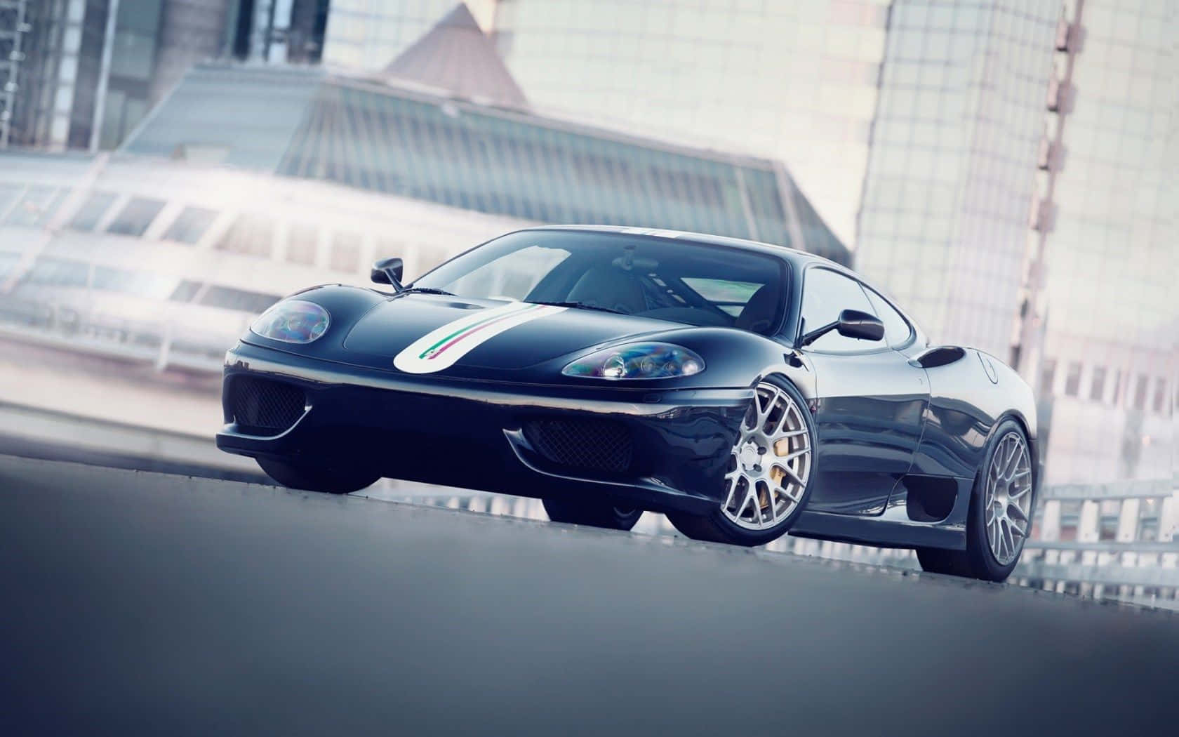 Ferrari 360 Modena 桌布 壁紙 — 一輛帶有義大利國旗條紋的時尚黑色法拉利跑車，在模糊的現代建築背景下，於城市街道上滑行。