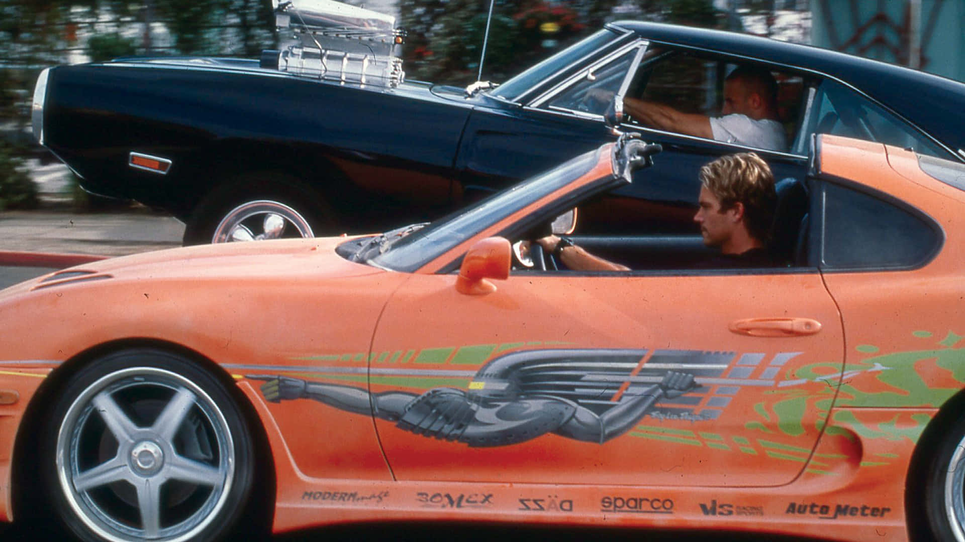 Fast And Furious 1 桌布 壁紙 — 一輛帶有翼形圖案的橙色改裝跑車在陽光明媚的街道上飛馳而過一輛黑色經典肌肉車。