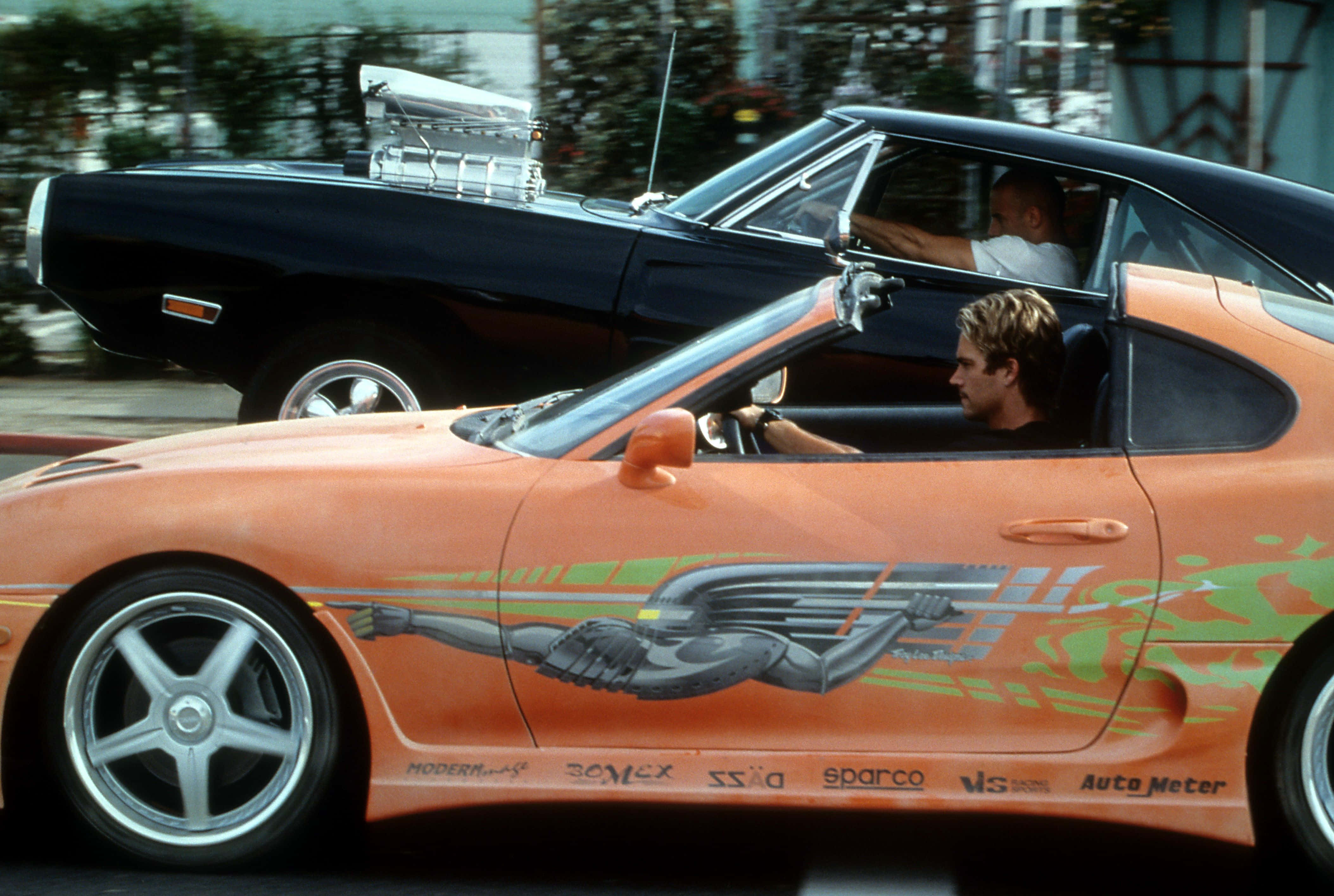 Fast And Furious 桌布 壁紙 — 一輛帶有翼狀圖案的橙色跑車在城巿街道上超越一輛黑色肌肉車。