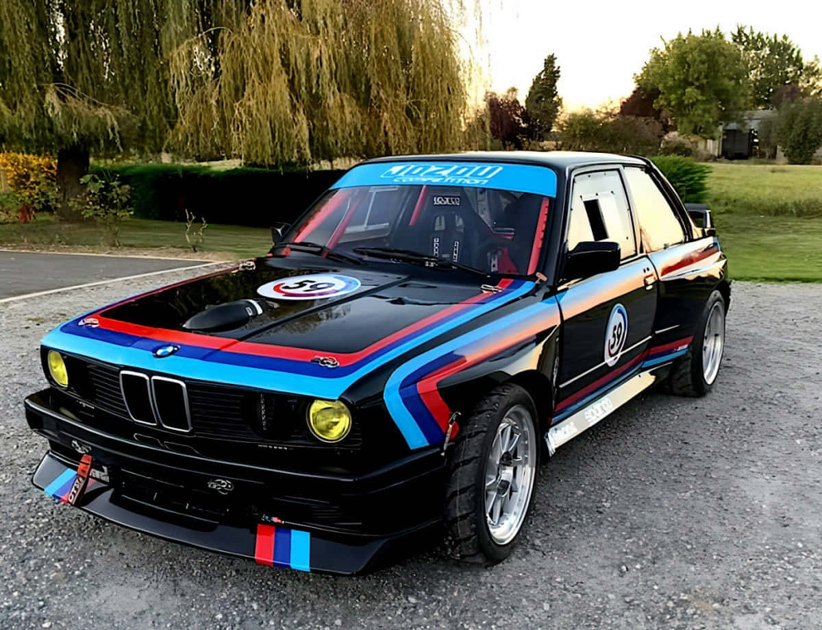 BMW 動圖 桌布 壁紙 — 一輛帶有藍、紅、白條紋的黑色寶馬賽車在黃金時刻停泊在垂柳附近的礫石上。