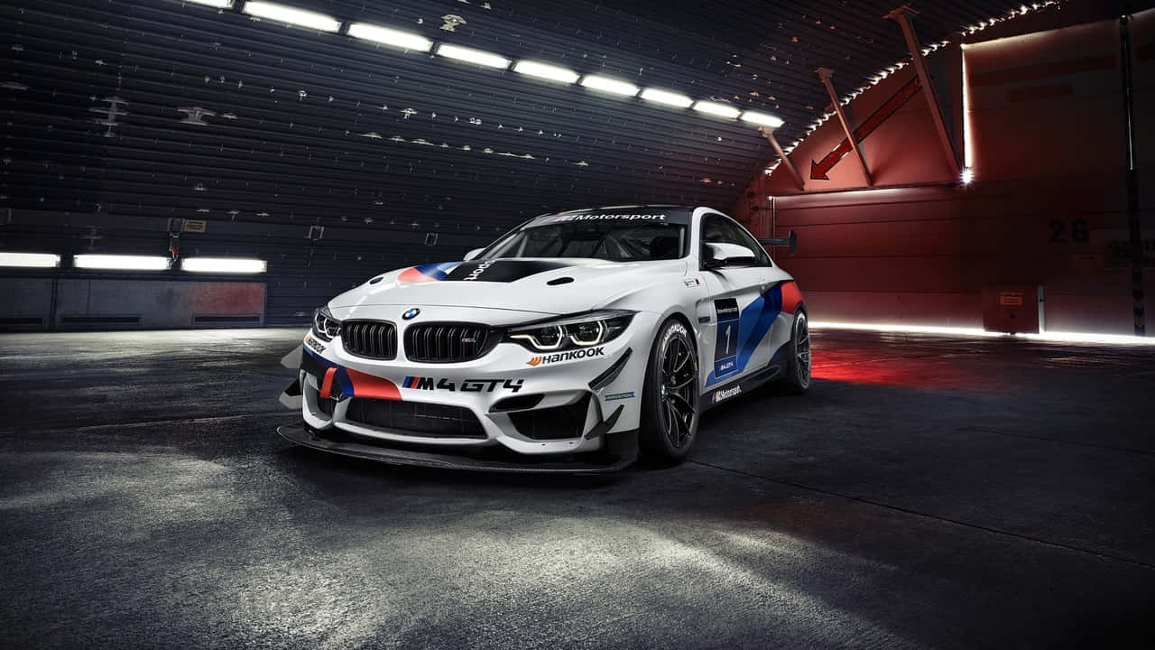 720p Bmw 桌布 壁紙 — 一輛帶有藍色、紅色和黑色塗裝的白色BMW M4 GT4賽車停放在昏暗的工業車庫中，在明亮的燈光下。