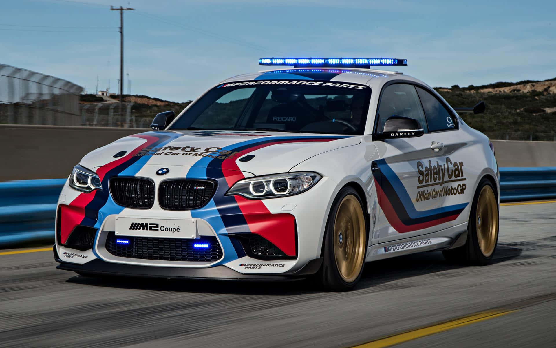 Safety Car 桌布 壁紙 — 一輛帶有藍色、紅色和黑色賽車條紋的白色BMW M2 Coupe安全車，在晴朗的藍天下沿著賽道飛馳。