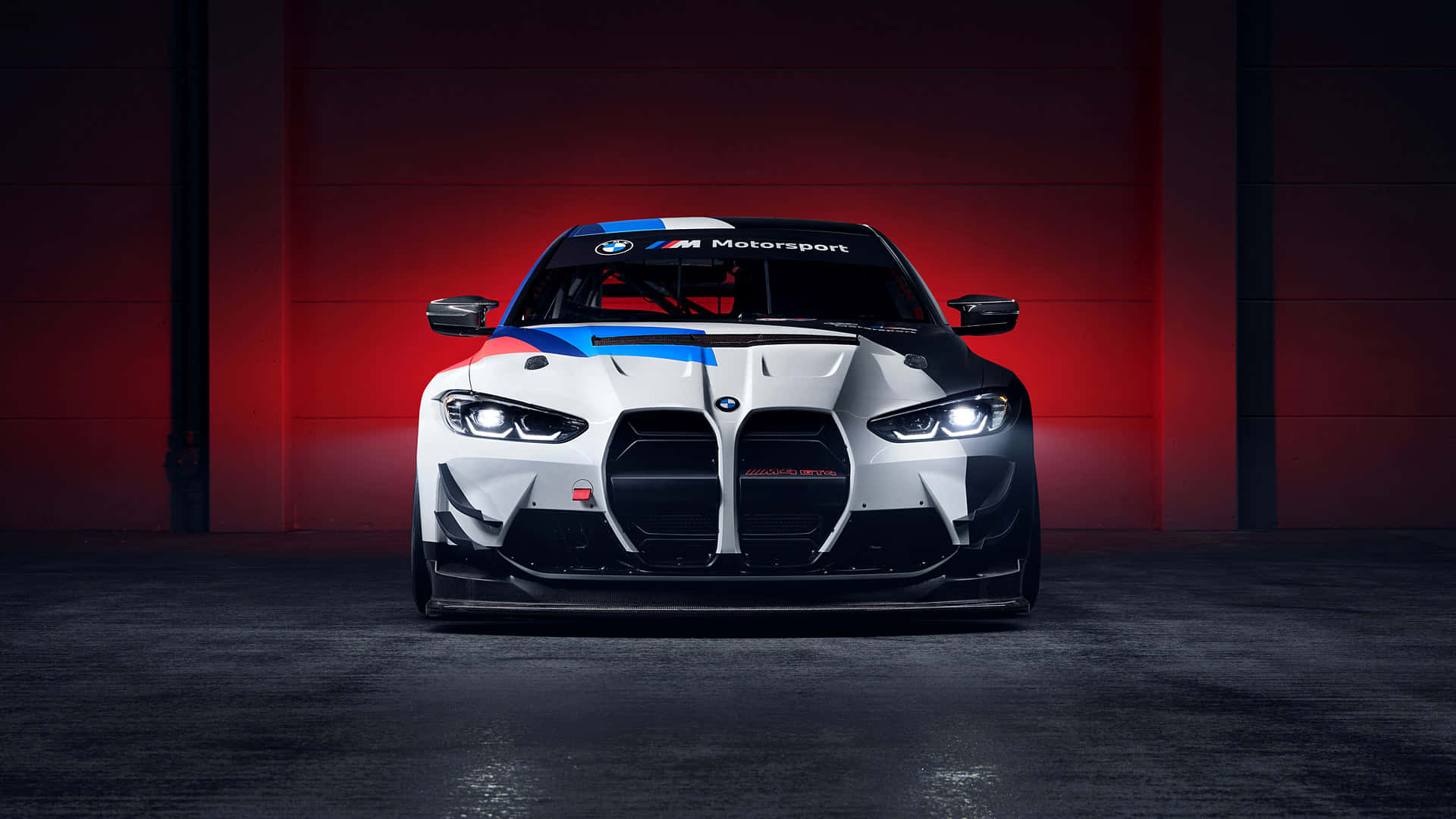 跑車 桌布 壁紙 — 一輛帶有藍色和紅色點綴的白色BMW M Motorsport賽車，在黑暗的車庫中，在戲劇性的紅色燈光下直視前方。