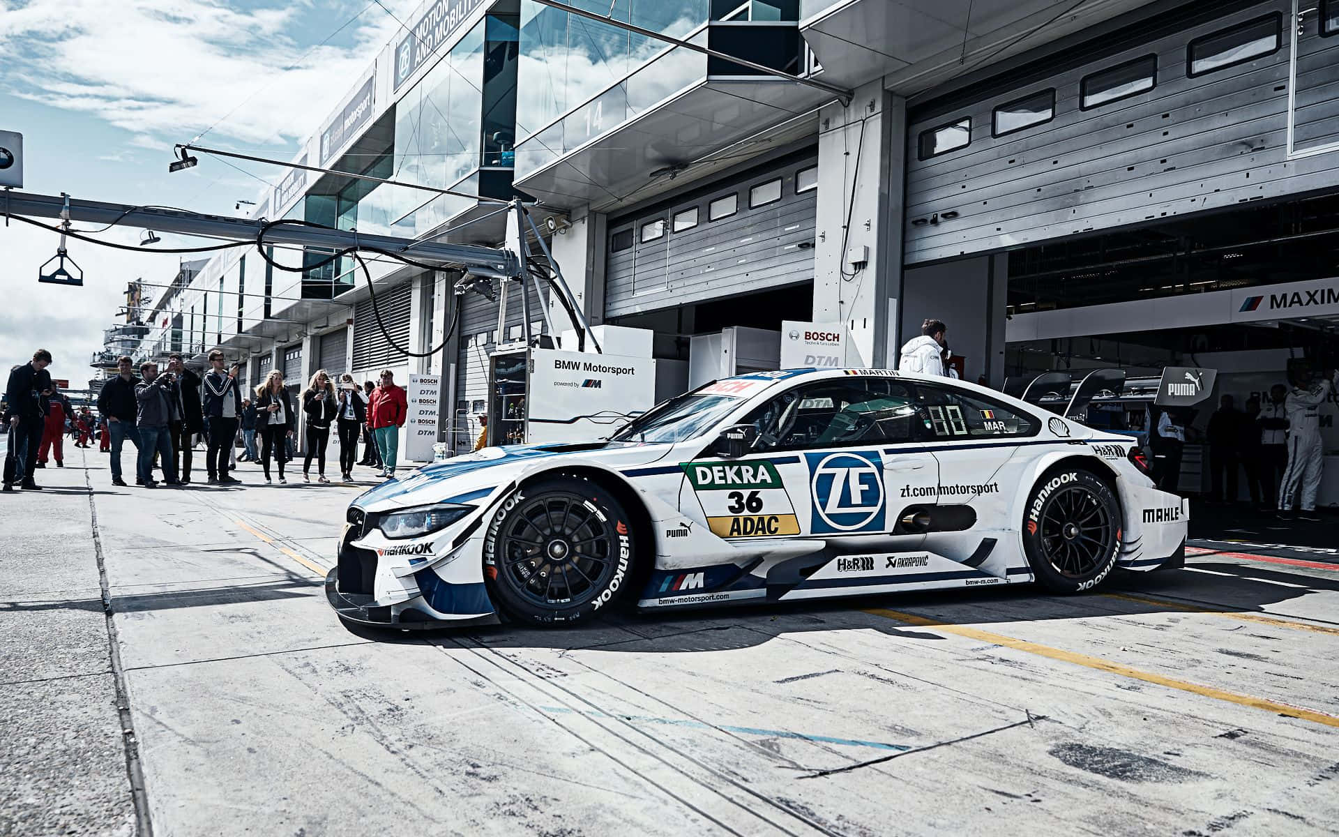 BMW 動圖 桌布 壁紙 — 一輛帶有藍色和黃色貼花的白色BMW賽車停在維修站車庫，觀眾在明亮的天空下觀看。