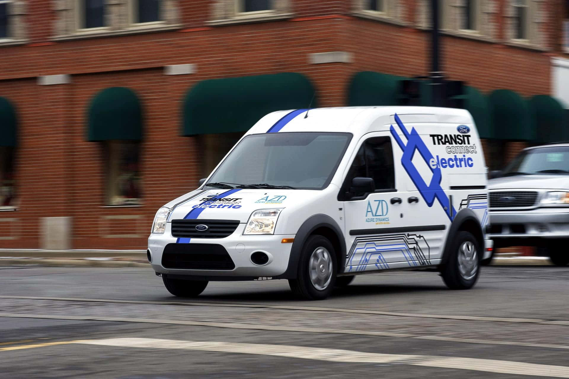 Ford Transit 桌布 壁紙 — 一輛帶有藍色圖案的白色福特 Transit Connect 電動貨車駛過城市街道，經過帶有綠色遮陽篷的磚砌建築。