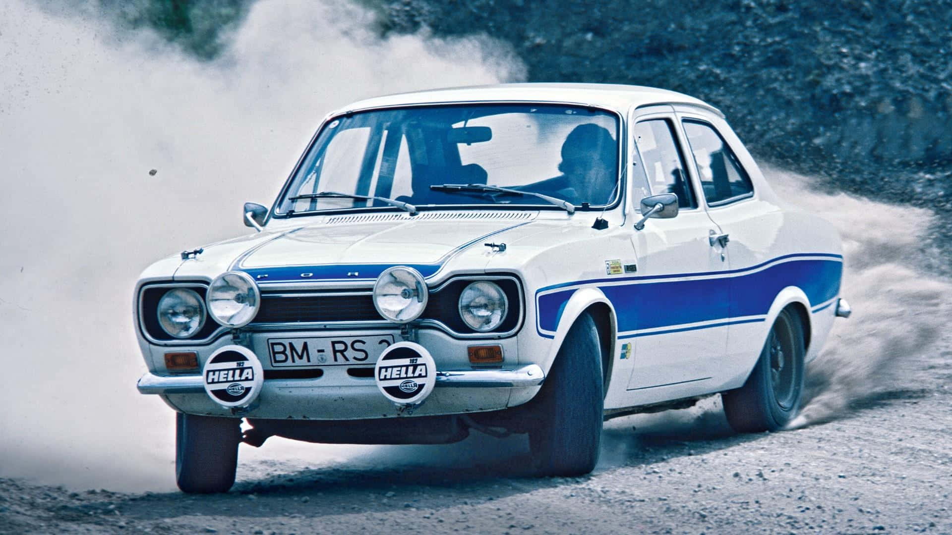 Ford Escort 桌布 壁紙 — 一輛帶有藍色條紋的白色福特Escort RS拉力賽車在崎嶇的泥路上疾馳，揚起碎石塵土。