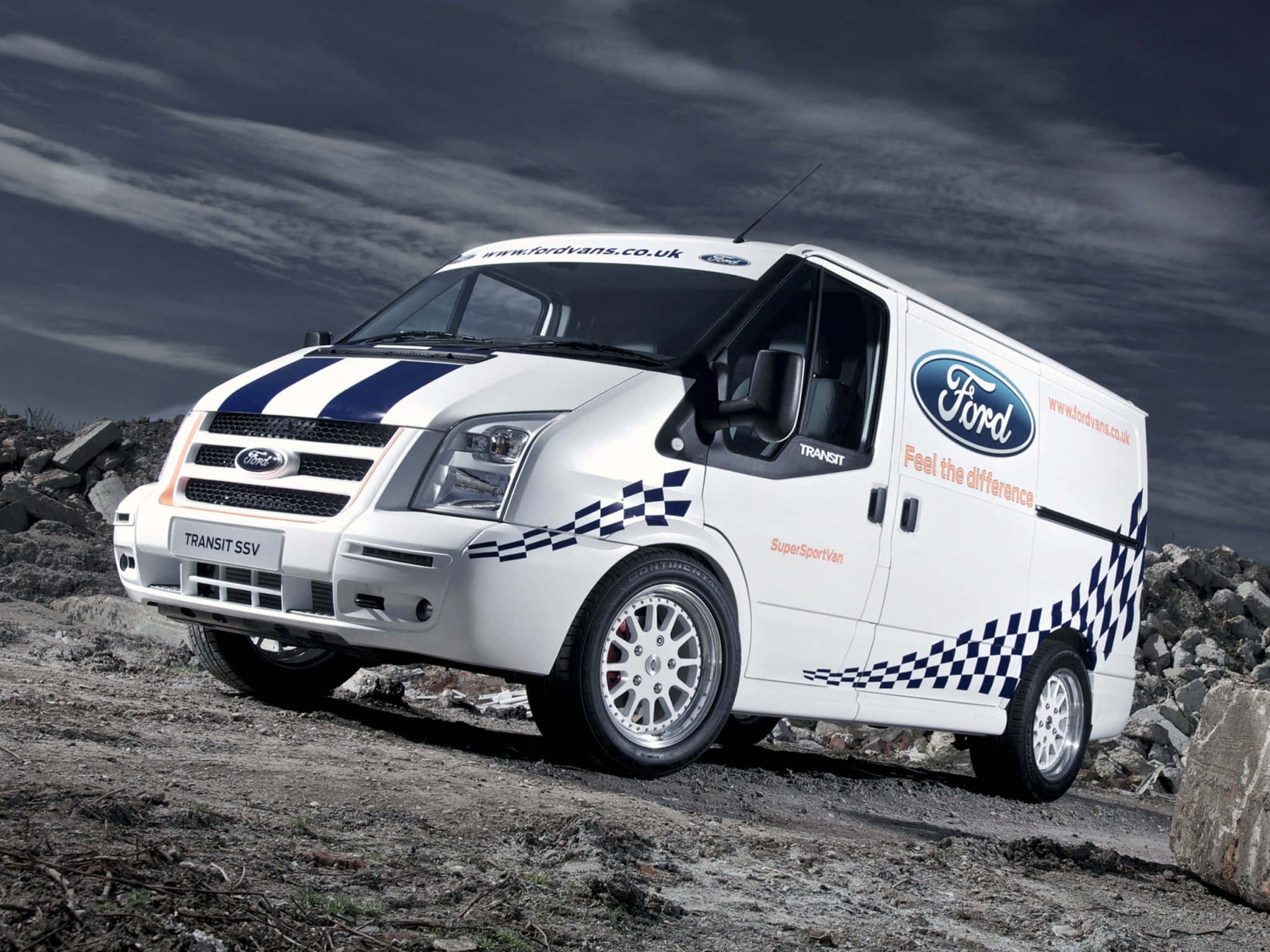 Ford Transit 桌布 壁紙 — 一輛帶有藍色賽車條紋和方格貼花的白色福特 Transit SSV 廂型車，站立在暴風雨天空下的岩石地形上。