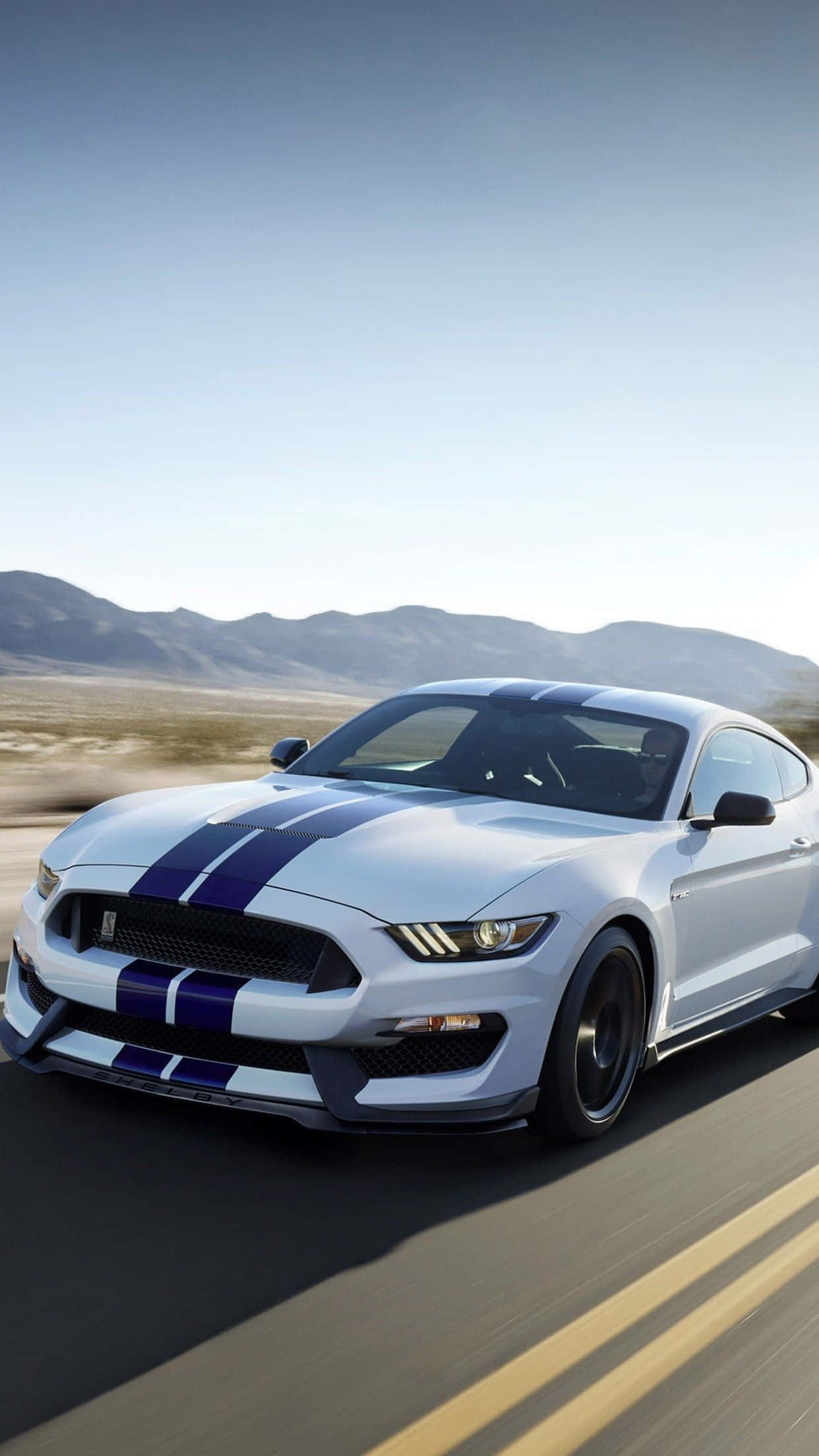 Mustang iPhone 桌布 壁紙 — 一輛帶有藍色賽車條紋的白色福特野馬在晴朗的藍天下沿著沙漠高速公路飛馳。