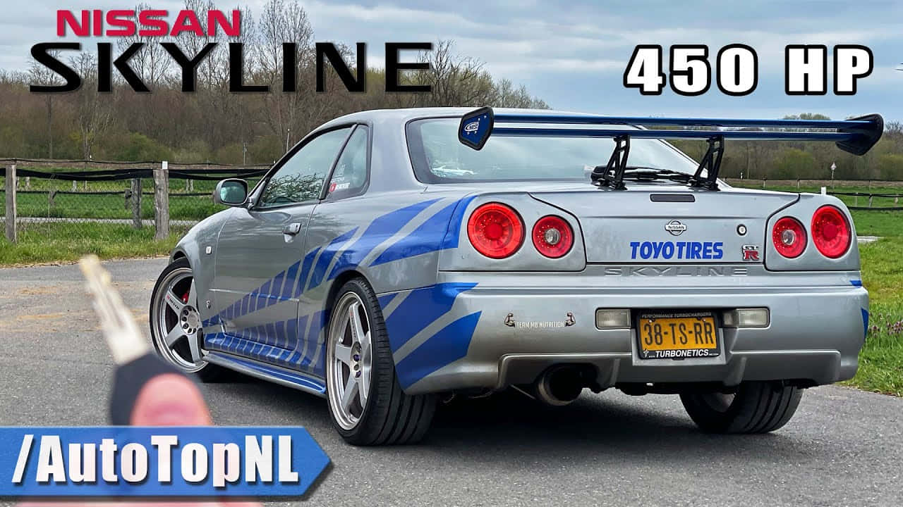 Paul Walker Skyline 桌布 壁紙 — 一輛帶有藍色賽車條紋的銀色日產Skyline停泊在碎石路上，背景有木柵欄和樹木。