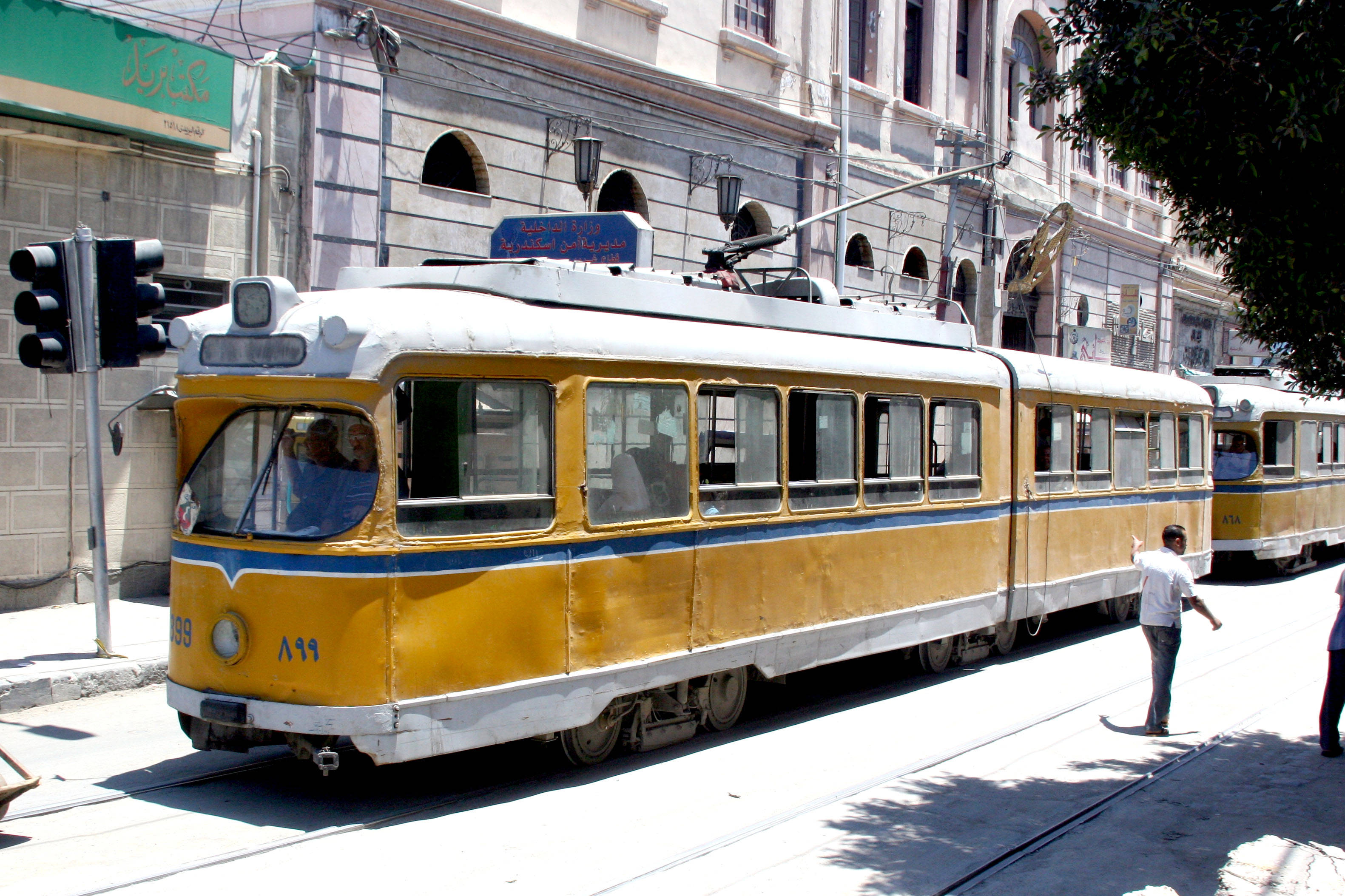 亞歷山卓 桌布 壁紙 — 一輛帶有藍色飾邊的復古黃色電車在陽光明媚的城市街道上滑行，經過歷史建築和行人。